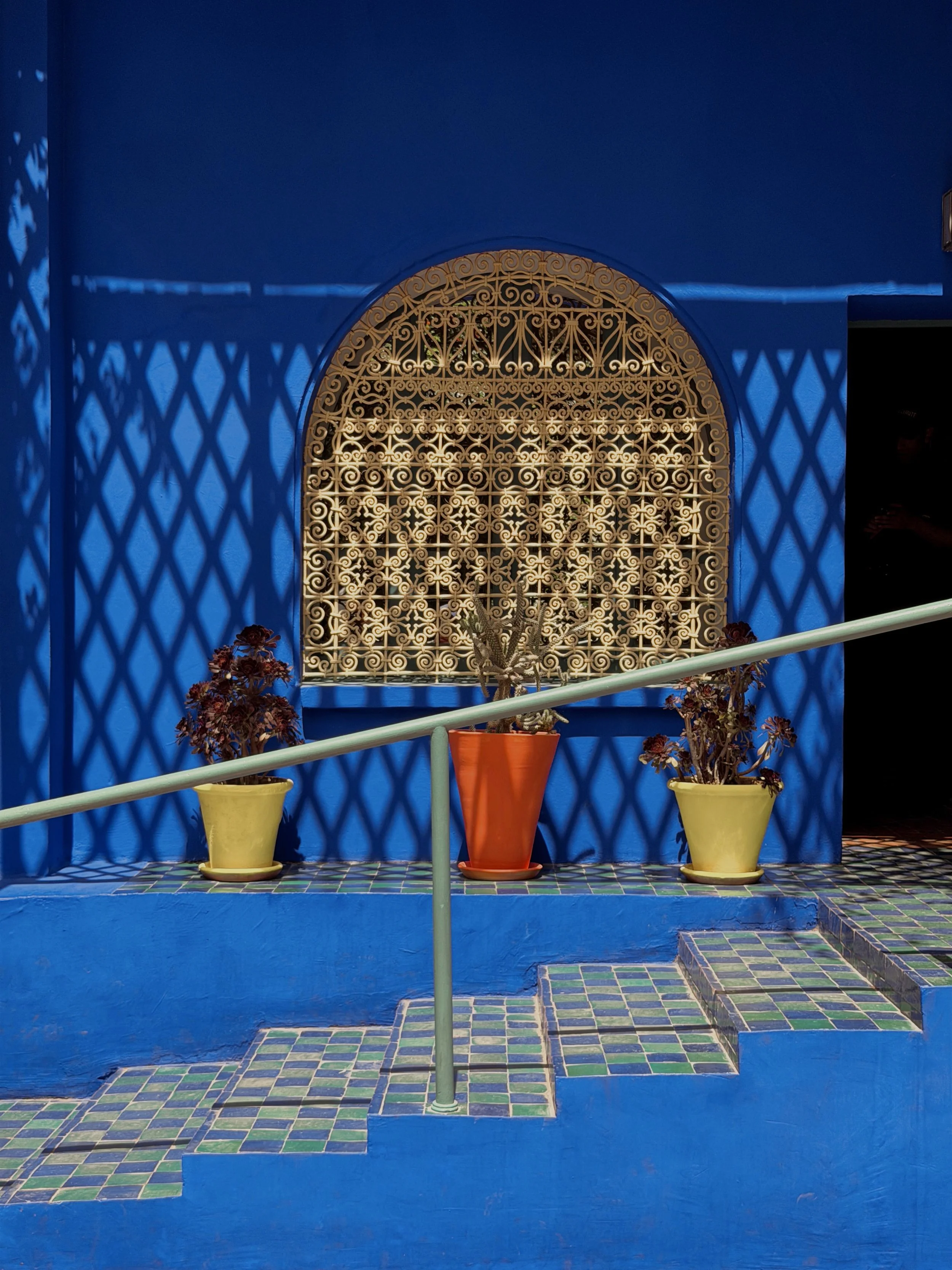 Cobalt-blue wall with a carved wooden window, yellow pots and steps in the foreground, Jardin Majorelle, Marrakech.