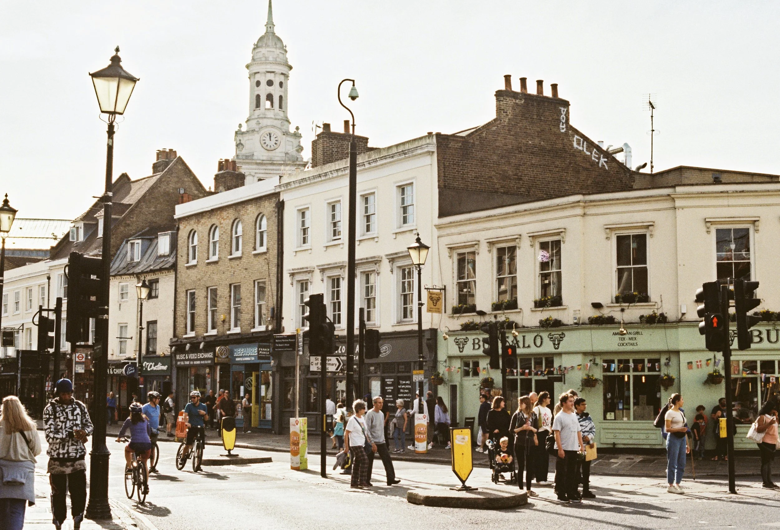 Busy street corner with a clock tower and pastel buildings in Greenwich, London.
