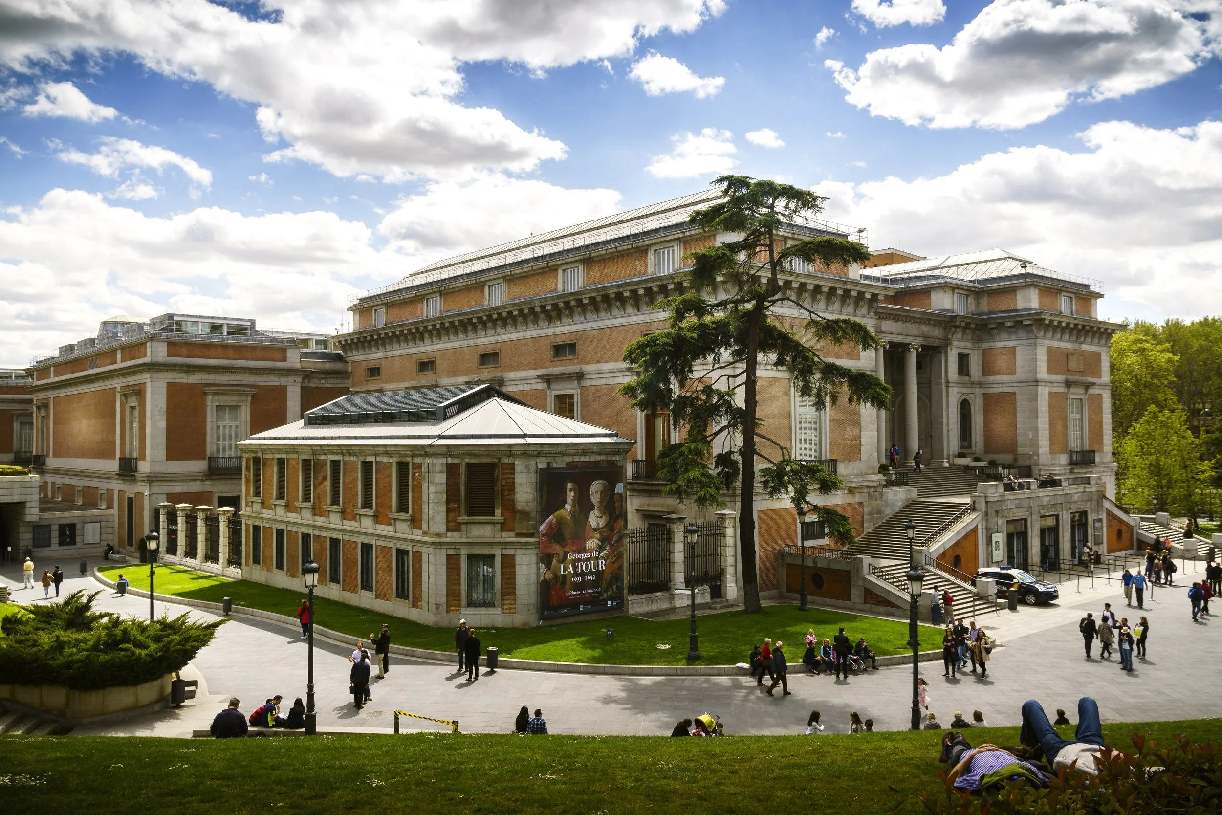 Museo del Prado brick and stone building with visitors on the lawn and steps under a partly cloudy sky, Madrid