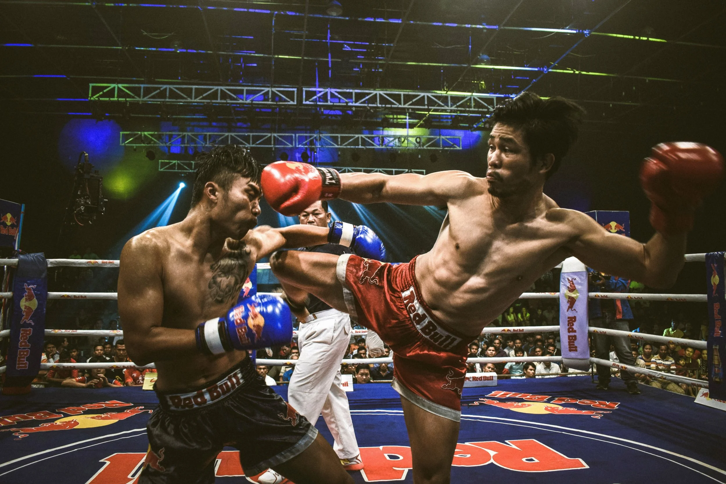 Two shirtless male Muay Thai fighters engaged in a fight in a ring with an audience watching. One fighter is throwing a kick while the other is defending with gloves.