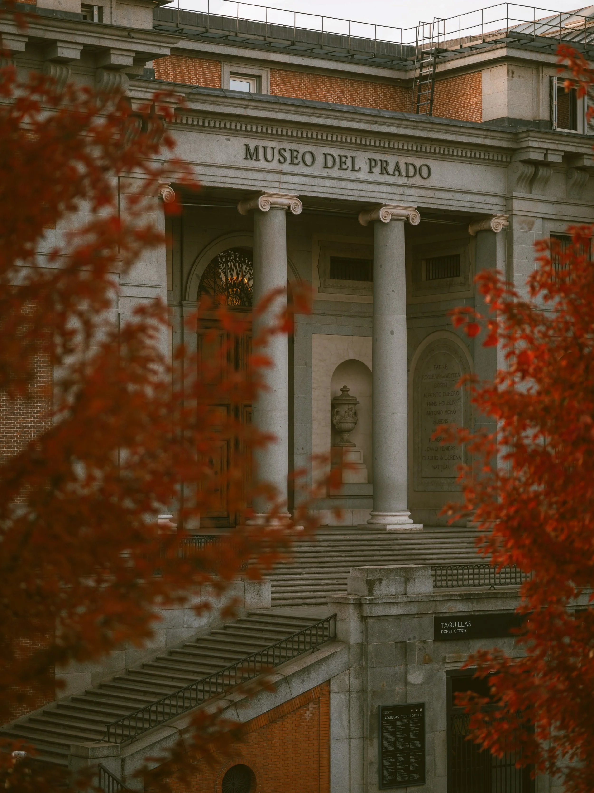 Facade of the Museo del Prado with stone columns and stairs behind red-leafed trees, Madrid