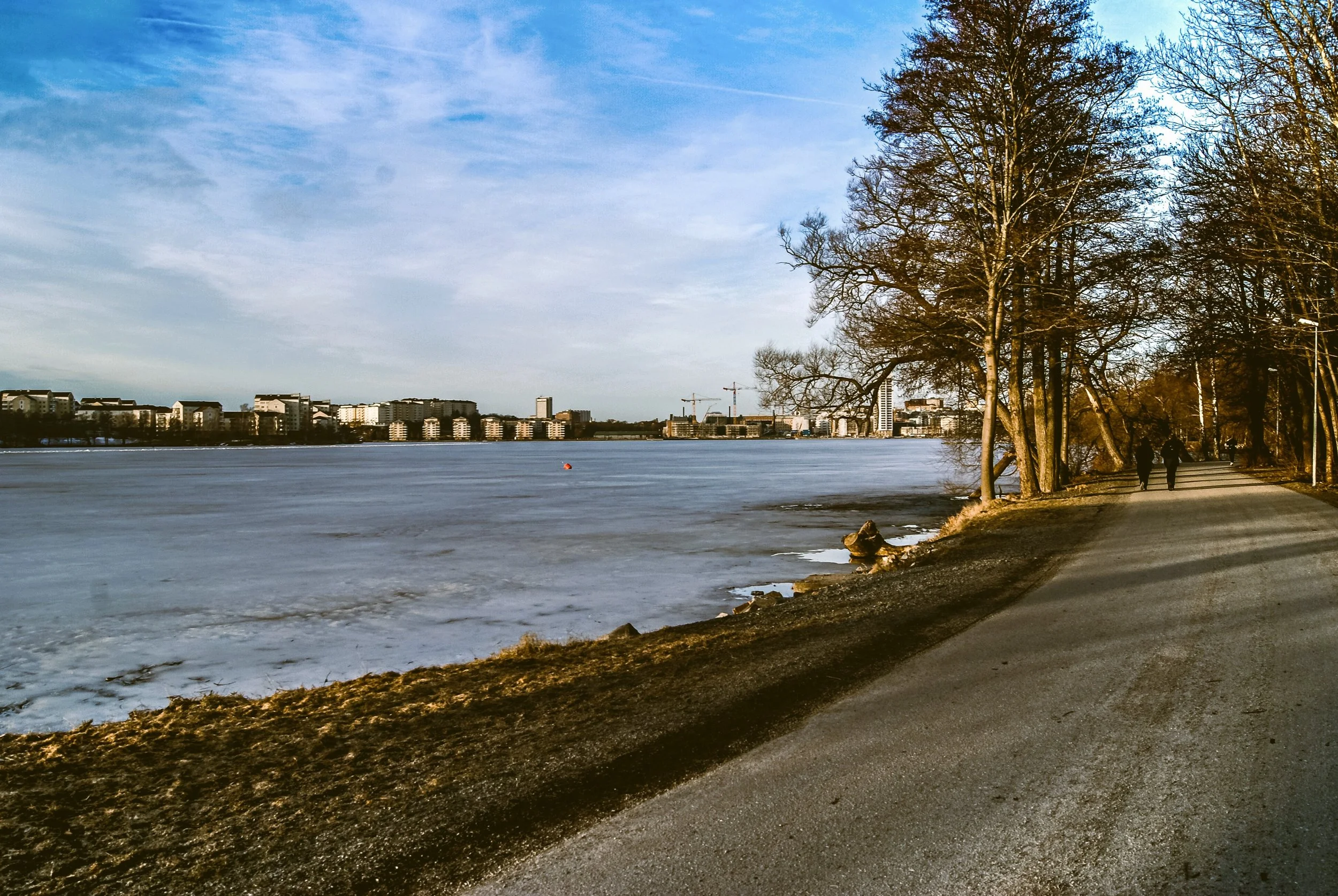 A winter scene by a frozen body of water with a city skyline in the background, trees on the right, and two people walking along the path on the right.