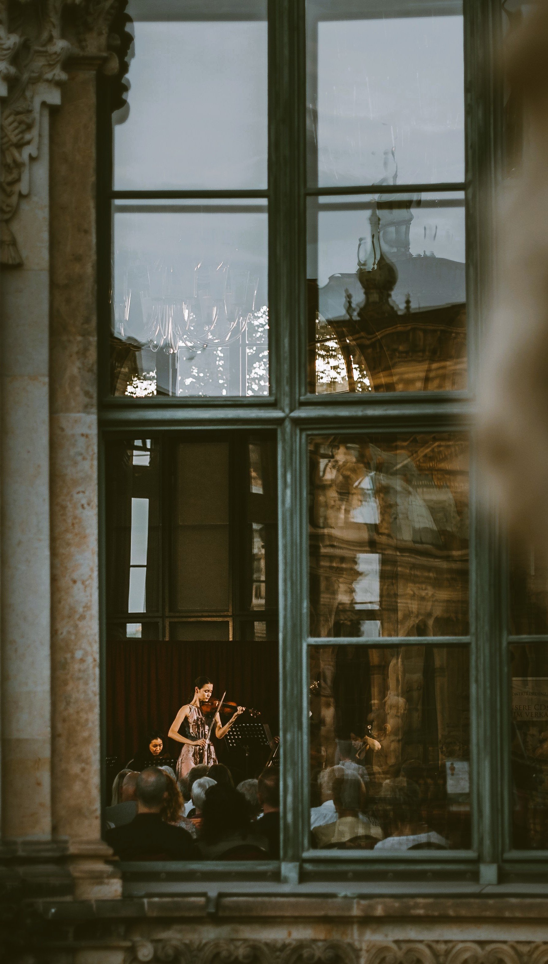 A woman playing the violin during a concert viewed through a large window with multiple panes, showing reflections and the interior chandelier.