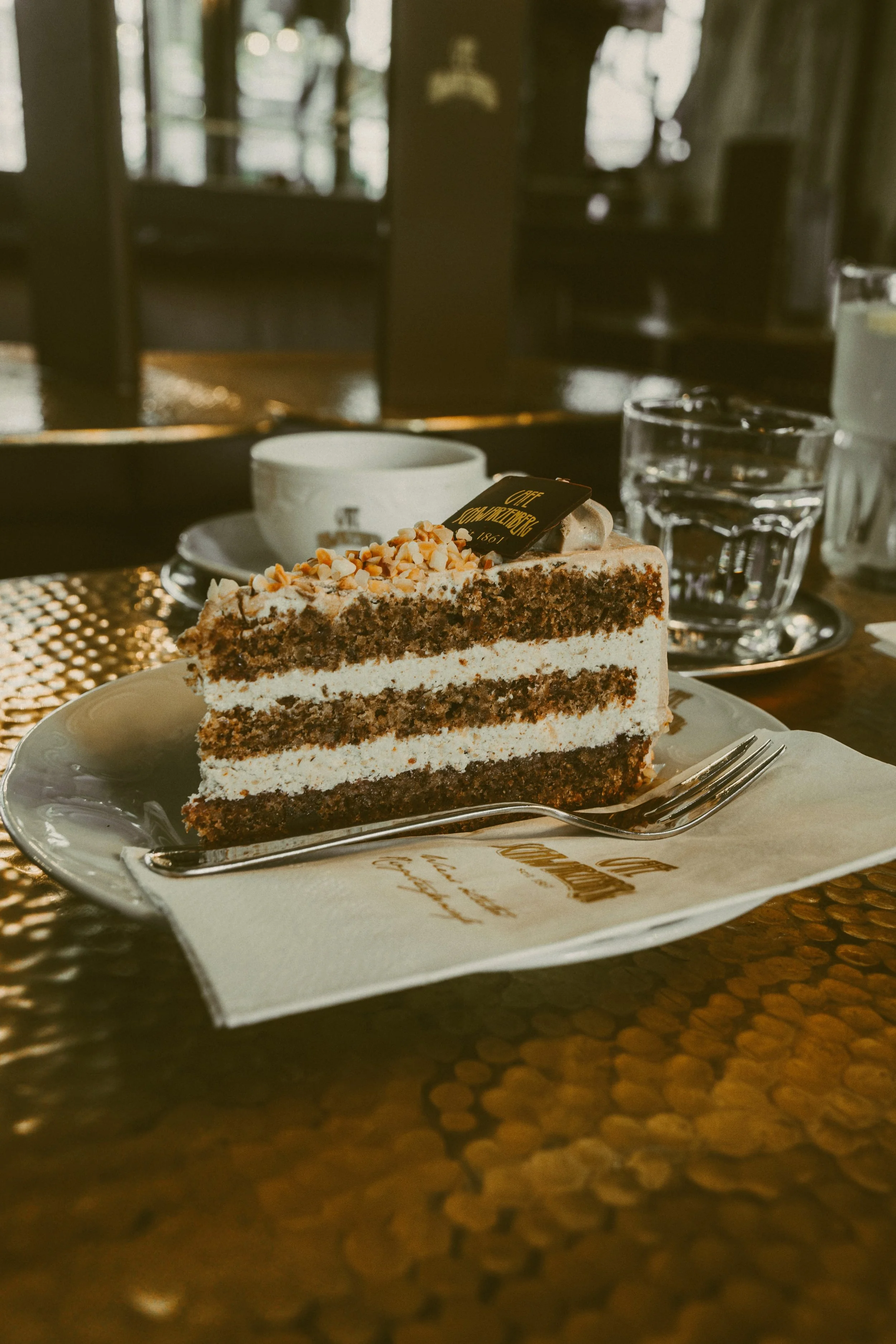 A slice of chocolate cake with white frosting, topped with chopped nuts, served on a white plate with a fork, set on a table in a cafe or restaurant.