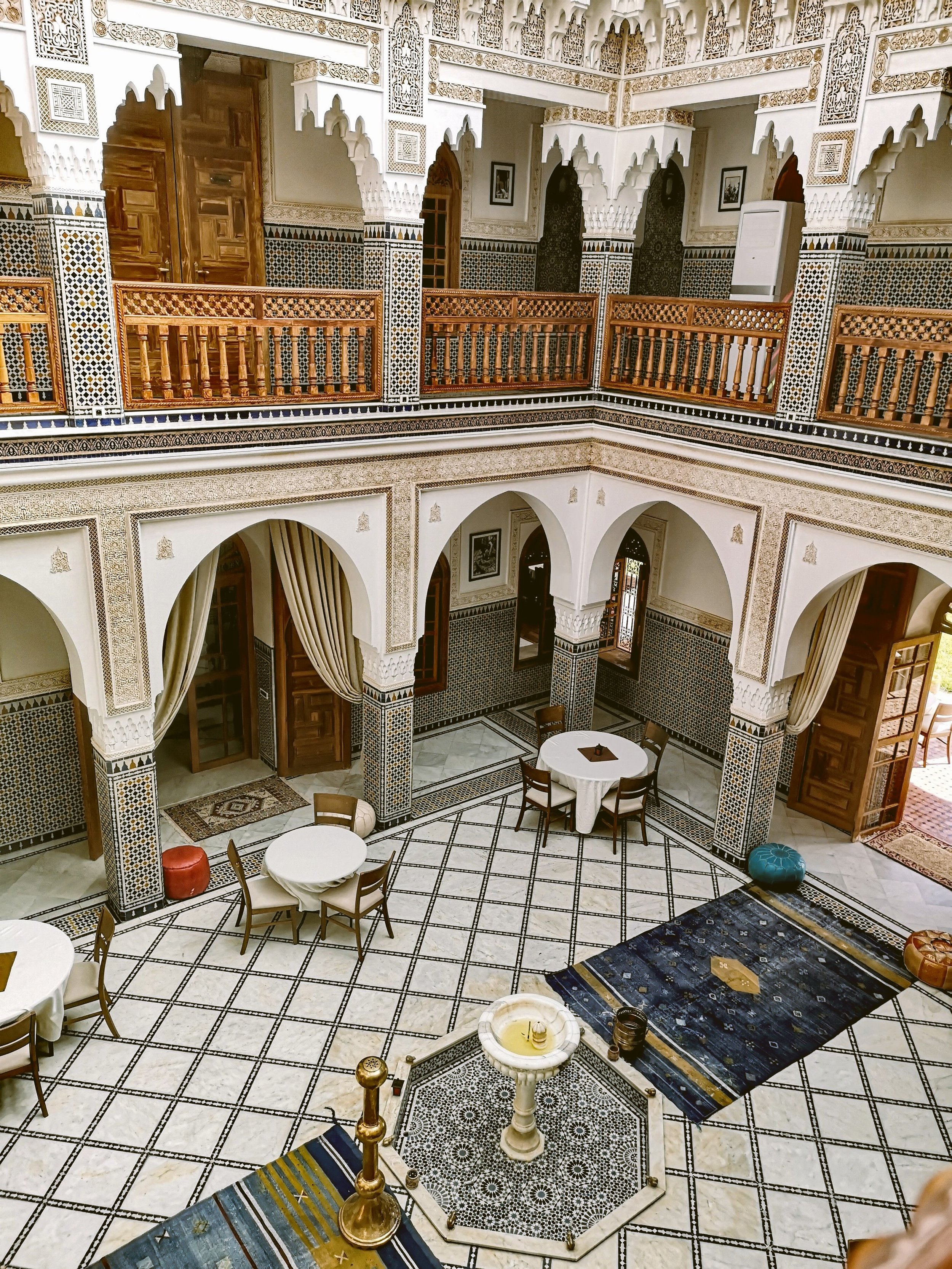 Riad courtyard seen from above with carved arches, upper galleries and lounge seating on a patterned tiled floor, Marrakech.