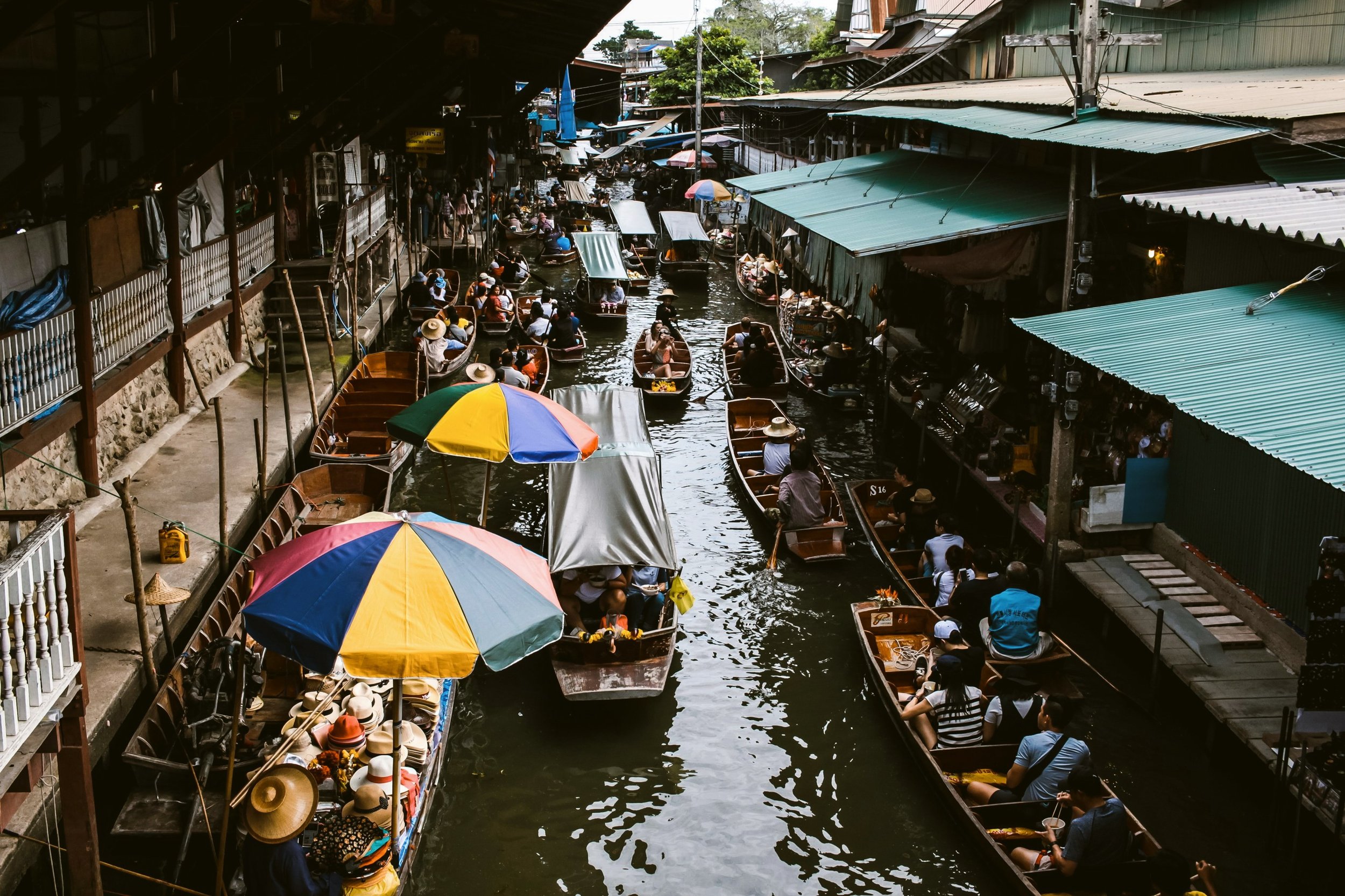 Boats with colorful umbrellas moving along a narrow canal lined with market stalls, Bangkok.