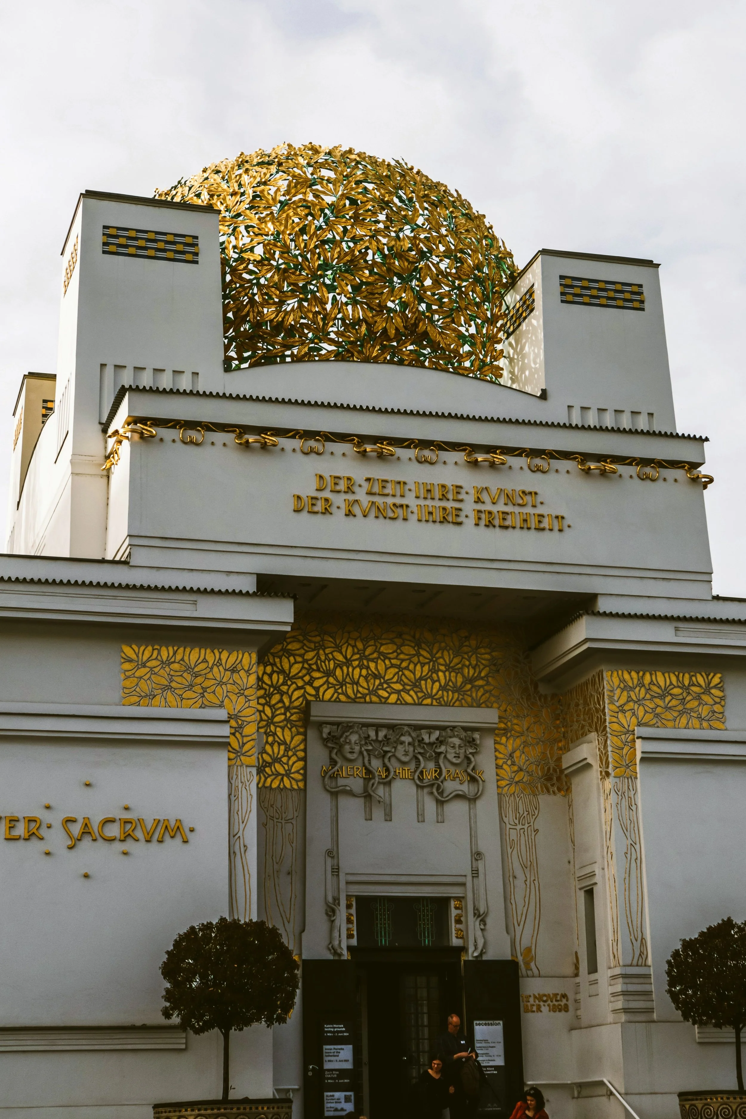 The image shows the exterior of the Secession building in Vienna, Austria, decorated with golden leaves and ornate white architectural details, including inscriptions in German.