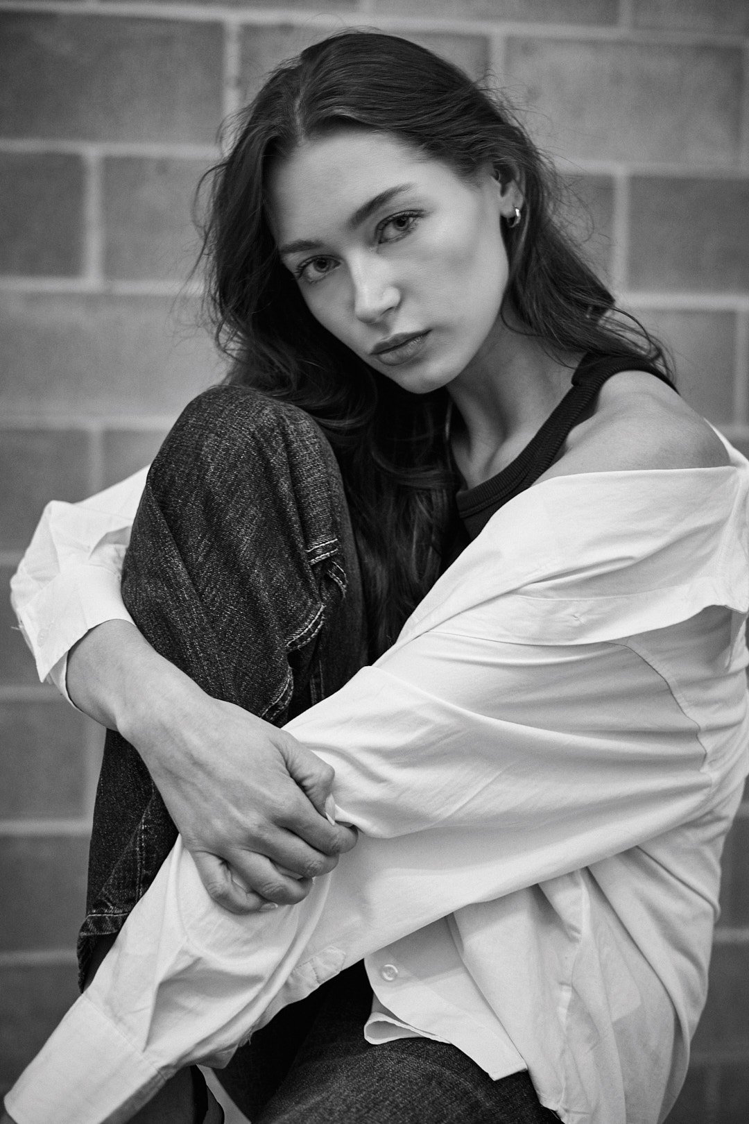 Retrato em preto e branco de uma mulher com cabelo longo, vestindo uma camiseta escura, um jeans e uma camisa branca, sentada contra uma parede de tijolos, olhando com uma expressão introspectiva para a câmera.