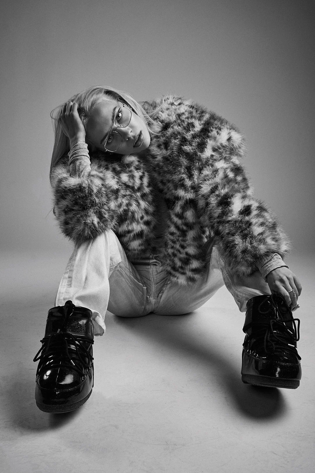 Imagem em preto e branco de uma mulher com cabelo loiro, óculos e expressão séria, usando casaco de pele animal, calça larga e botas foscas, sentada com as pernas cruzadas e apoiando a cabeça com uma mão.