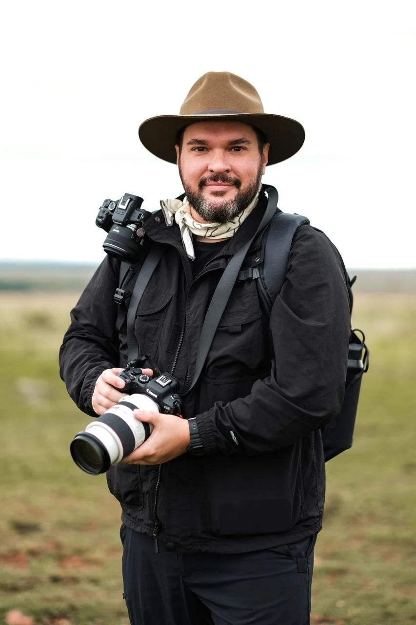 Homem fotógrafo com roupas de aventura, chapéu, mochila, segurando uma câmera profissional, em campo aberto ao ar livre.