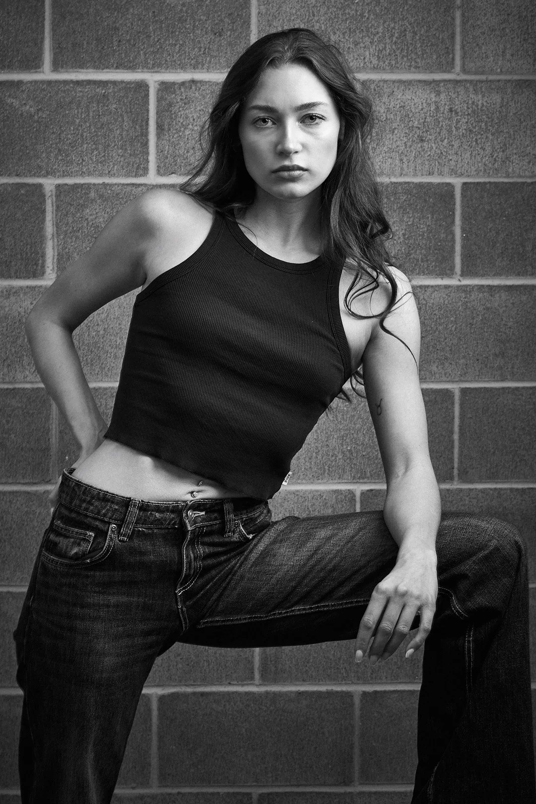 Mulher jovem com cabelos longos e escuros, de expressão séria, usando camiseta regata preta e jeans, posando contra uma parede de blocos de cimento.