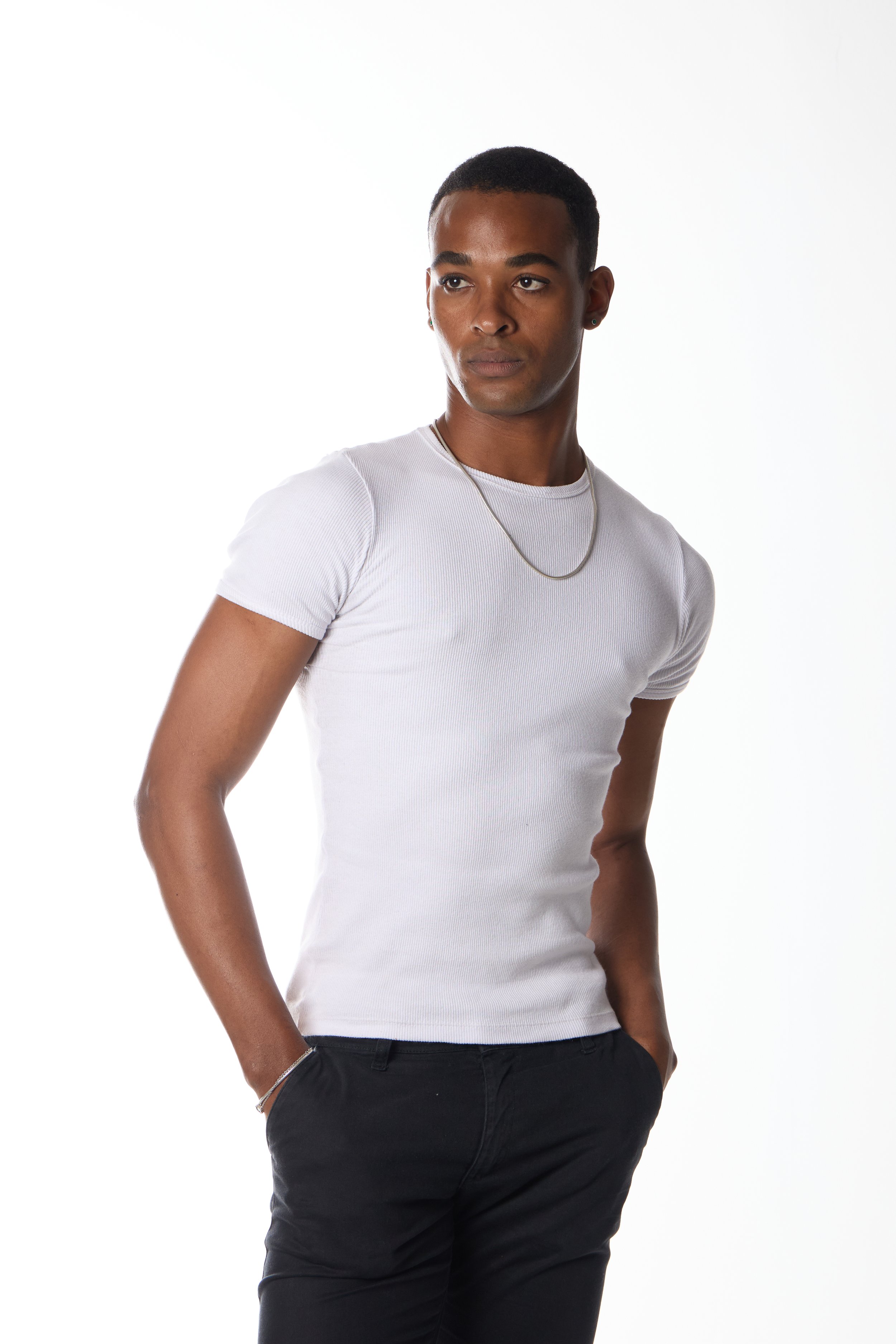 Pessoa jovem com pele morena clara, cabelo curto e escuro, usando camiseta branca justa, calça preta, brincos, uma corrente e uma pulseira, posando com as mãos no bolso contra fundo branco.