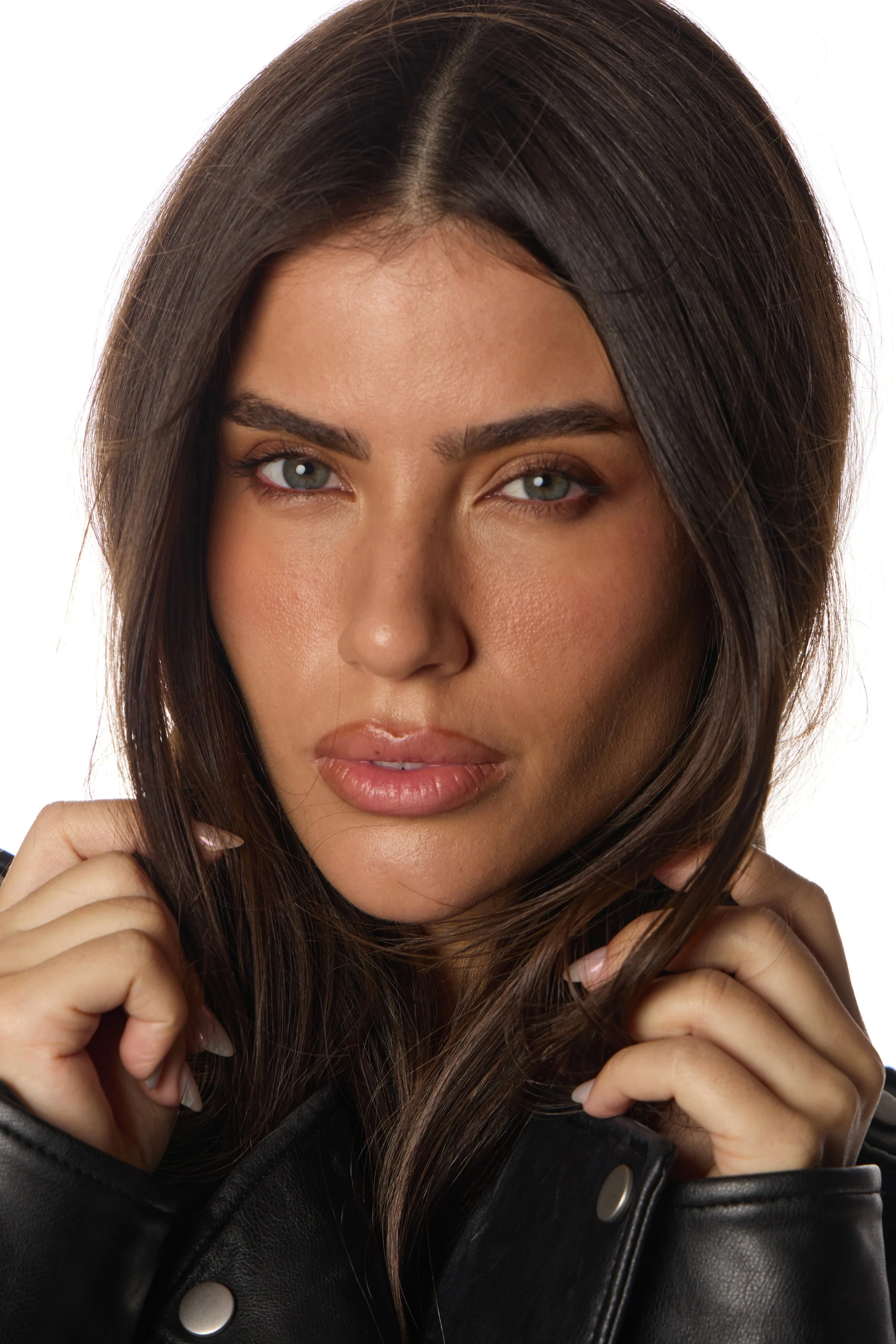 Mulher com cabelo castanho escuro, pele clara e olhos claros, usando jaqueta de couro preta, segurando o cabelo com as mãos.