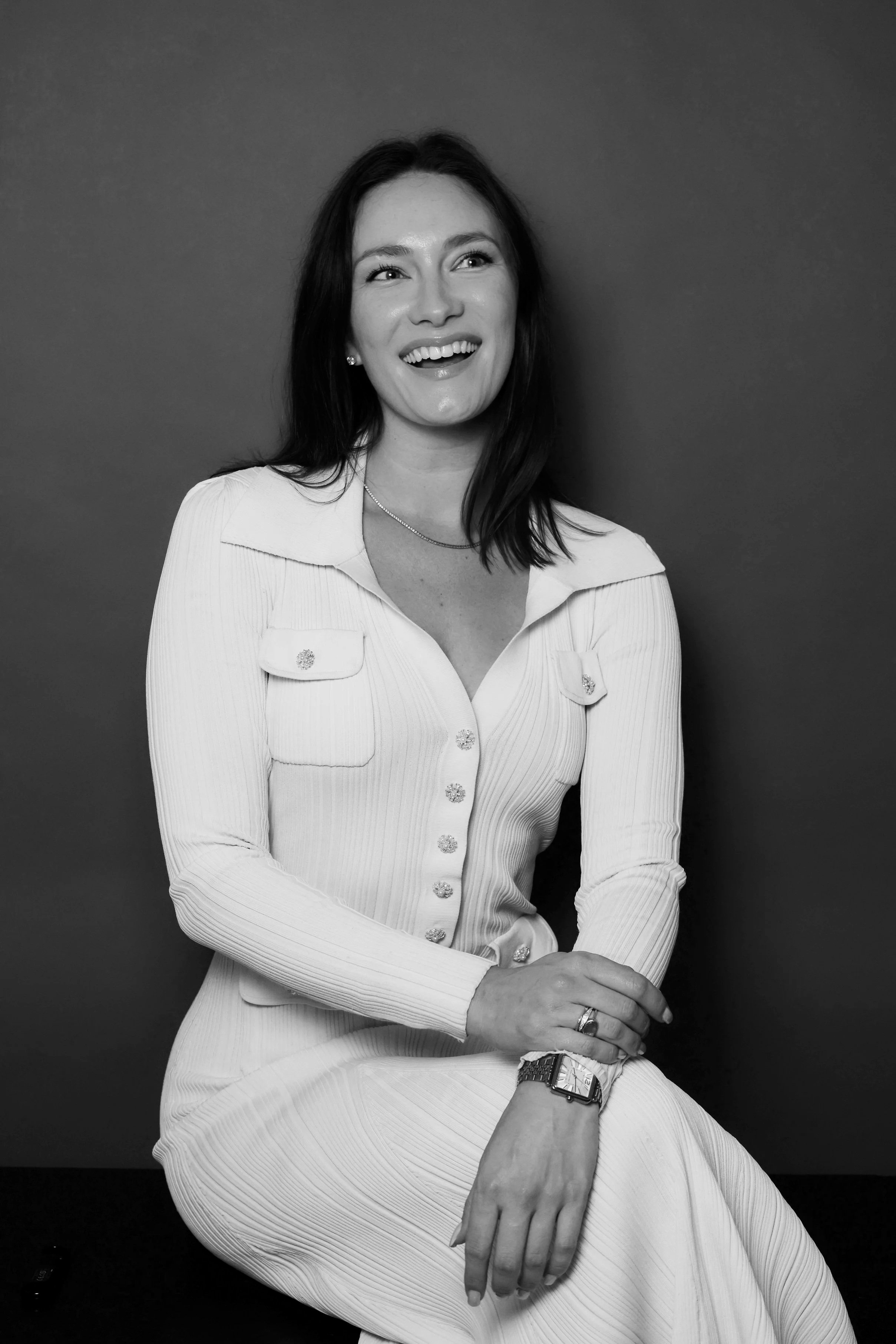 Black and white photograph of a woman with dark hair, smiling, wearing a long-sleeved, button-up dress with pockets, earrings, a necklace, a watch, and a ring, seated against a plain dark background.