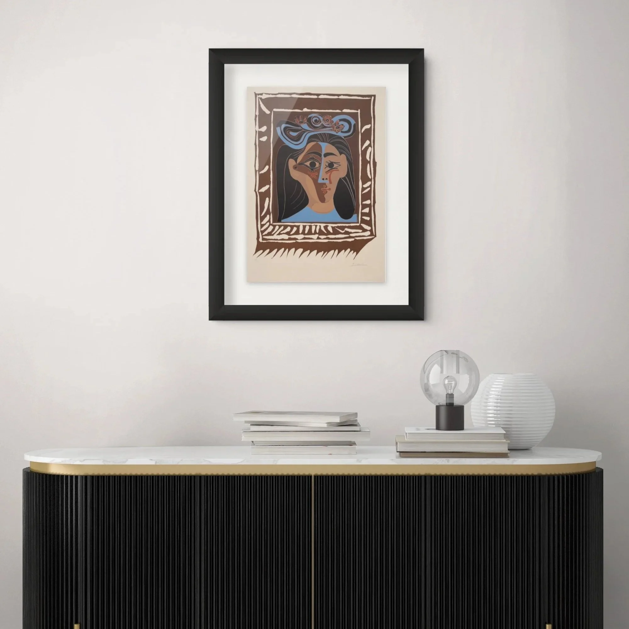 Modern interior decor with a framed abstract portrait of a woman, a table with a stack of books, a table lamp, and a small decorative object on a white wall.