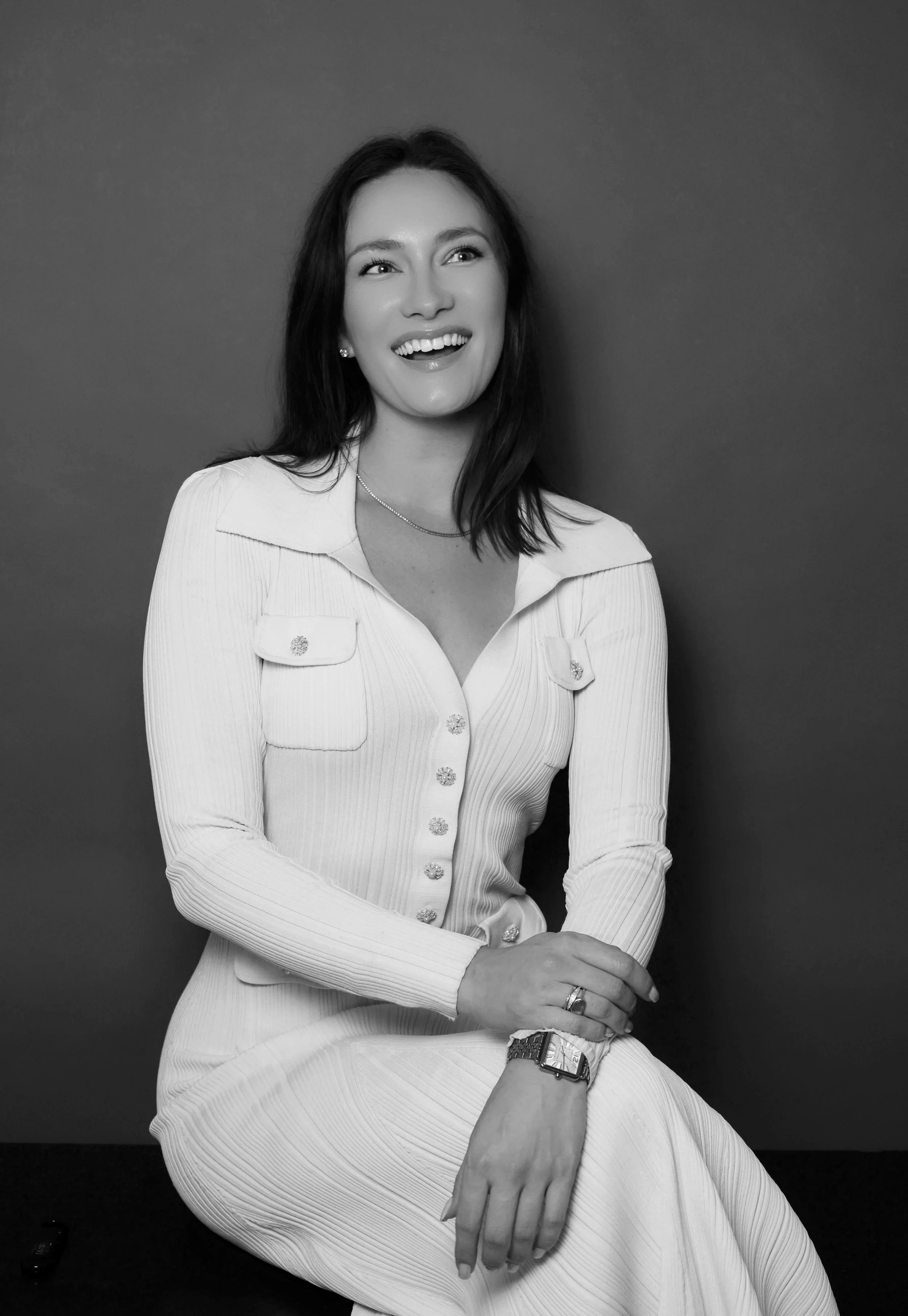 Black and white photo of a smiling woman with dark hair, wearing a white long-sleeved dress with buttons and pockets, sitting against a plain dark background.