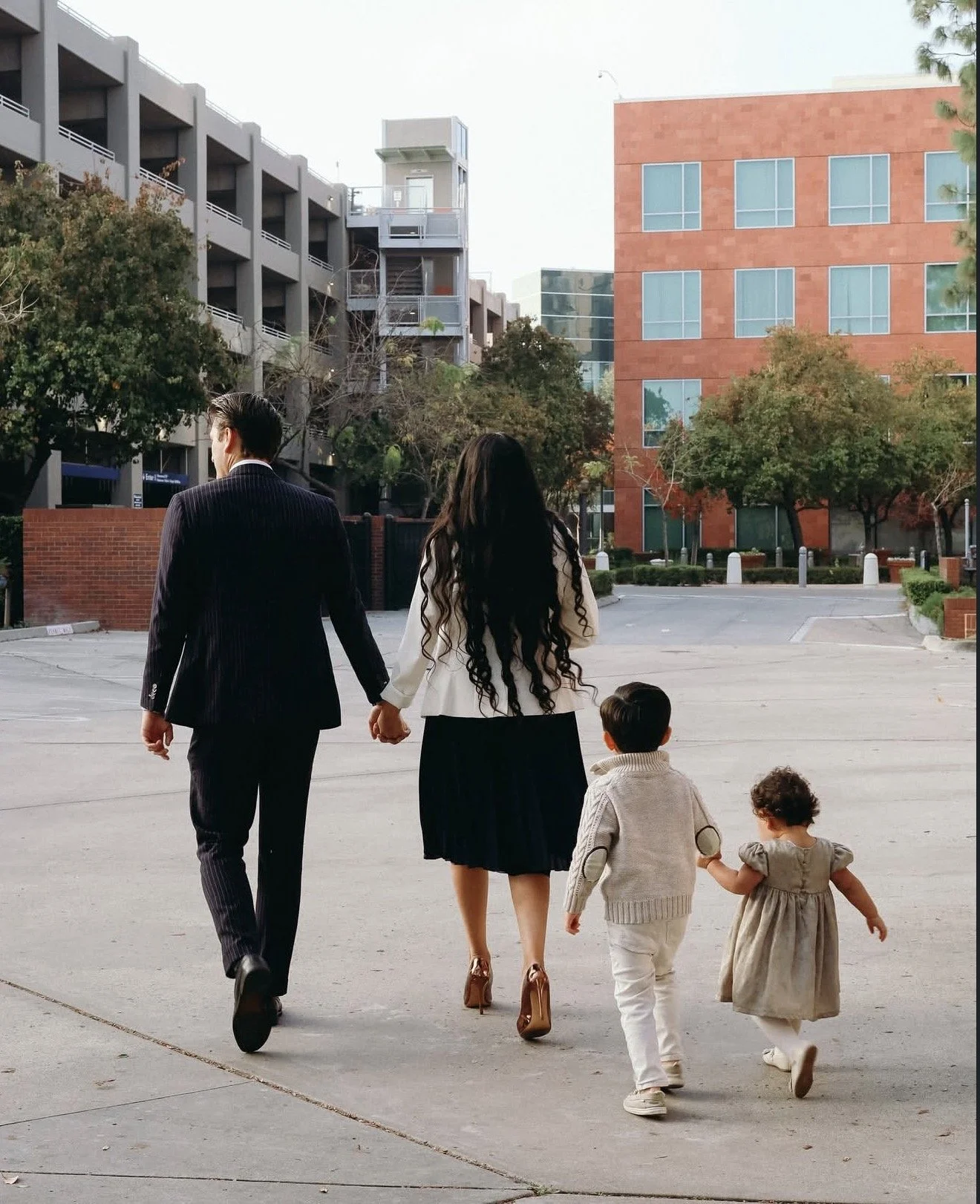 A family of five holding hands and walking in an urban parking lot with buildings and trees in the background.