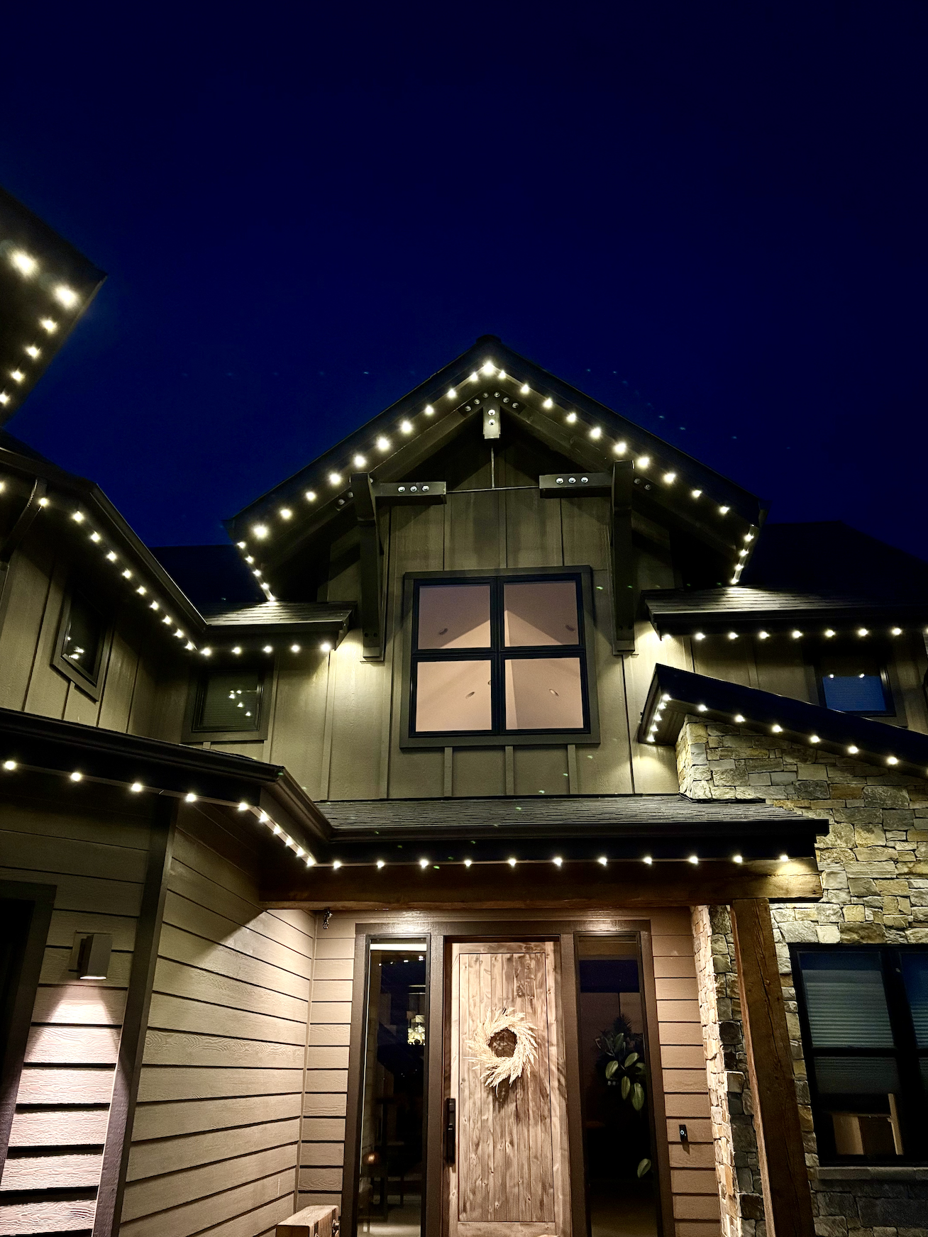 Exterior of a house at night decorated with string lights around eaves and roofline, featuring a wooden door with a wreath, and various windows.
