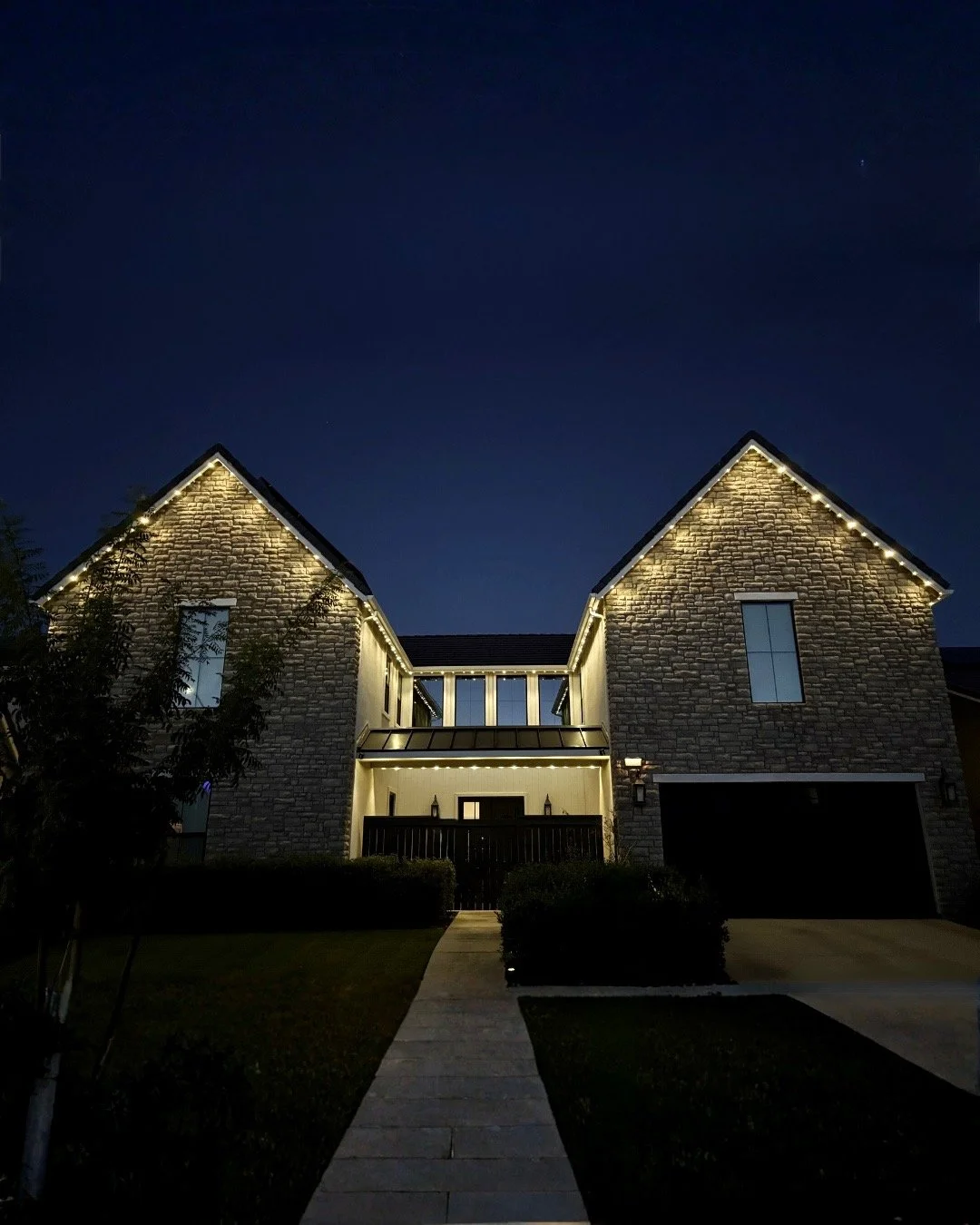 Nighttime view of a two-story house with stone exterior, decorated with string lights along the rooflines and balcony, with a walkway leading to the front door, shrubs, and a tree in the front yard.