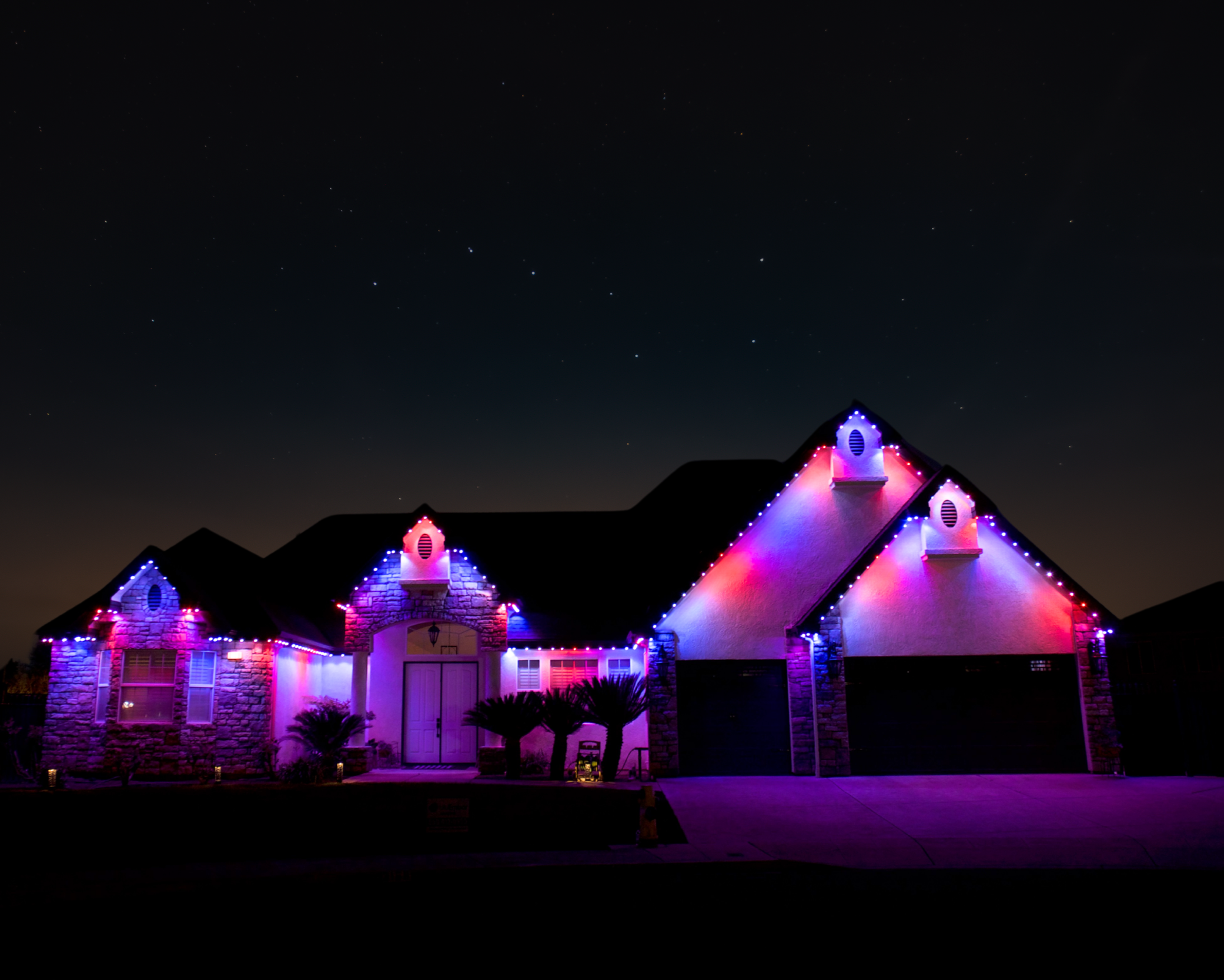 A house decorated with colorful Christmas lights at night. The house has a peaked roof, stone and stucco exterior, and trees in the front yard. The sky above is dark with visible stars.
