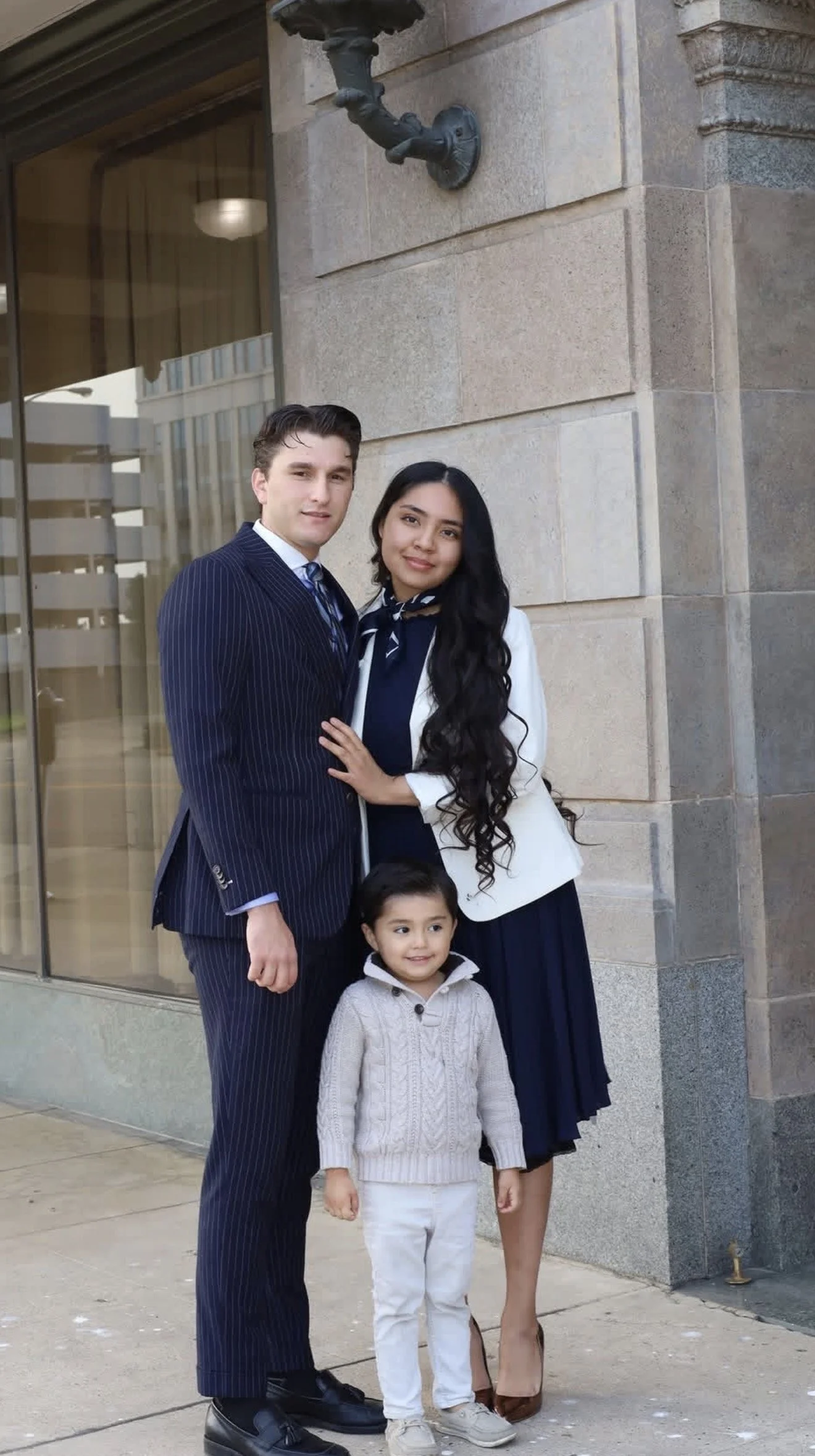 A family of four standing on a sidewalk in front of a building with large glass windows and stone walls. The father is wearing a pinstripe suit, the mother is dressed in a white blazer and navy dress, and two children are with them, a girl in a knit sweater, and a young boy in a light-colored sweater.