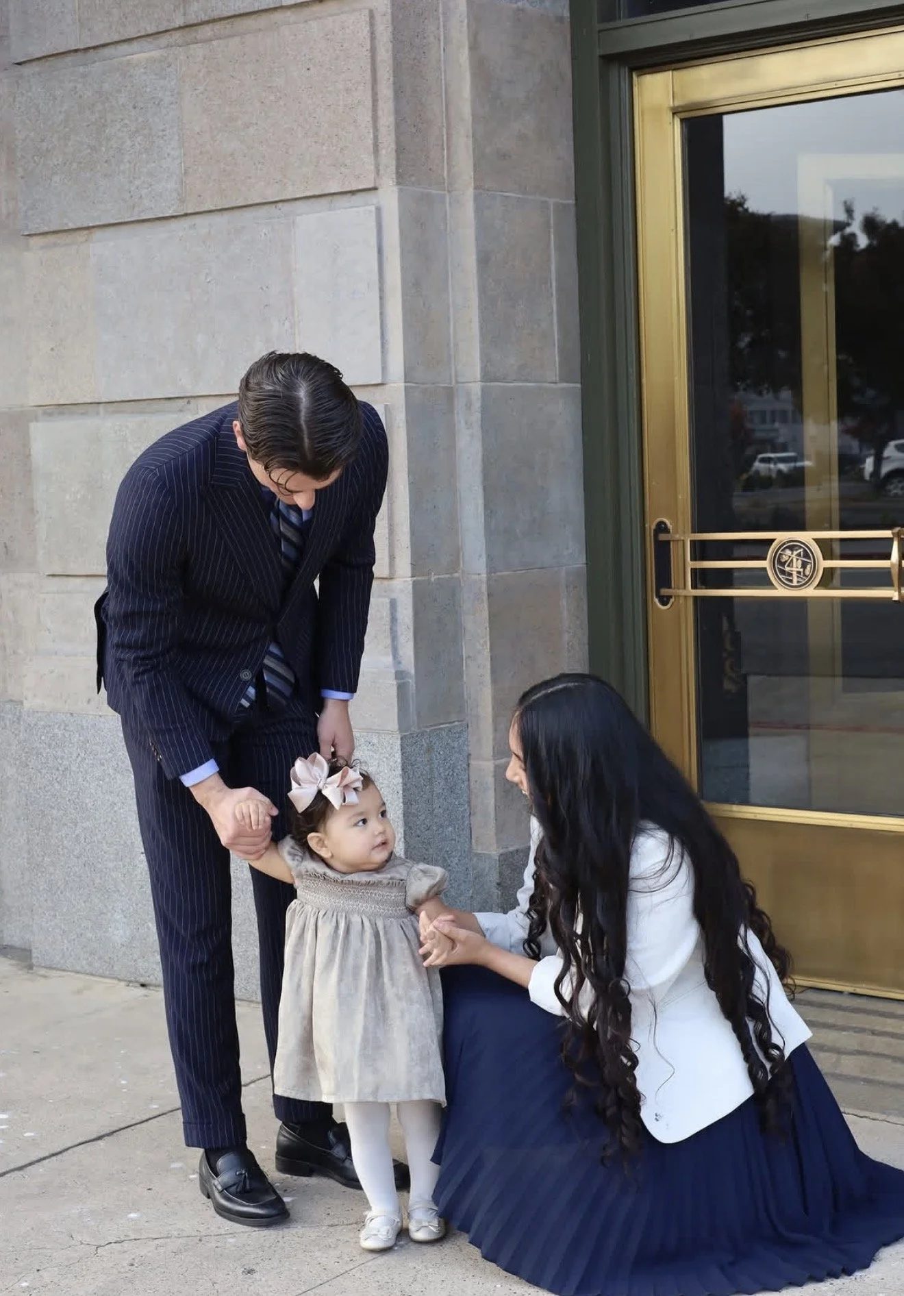 A man in a pinstripe suit and a woman with long dark hair are helping a young girl with a big bow in her hair. They are outside a building with a stone wall and a gold-colored door, possibly during a formal event or celebration.