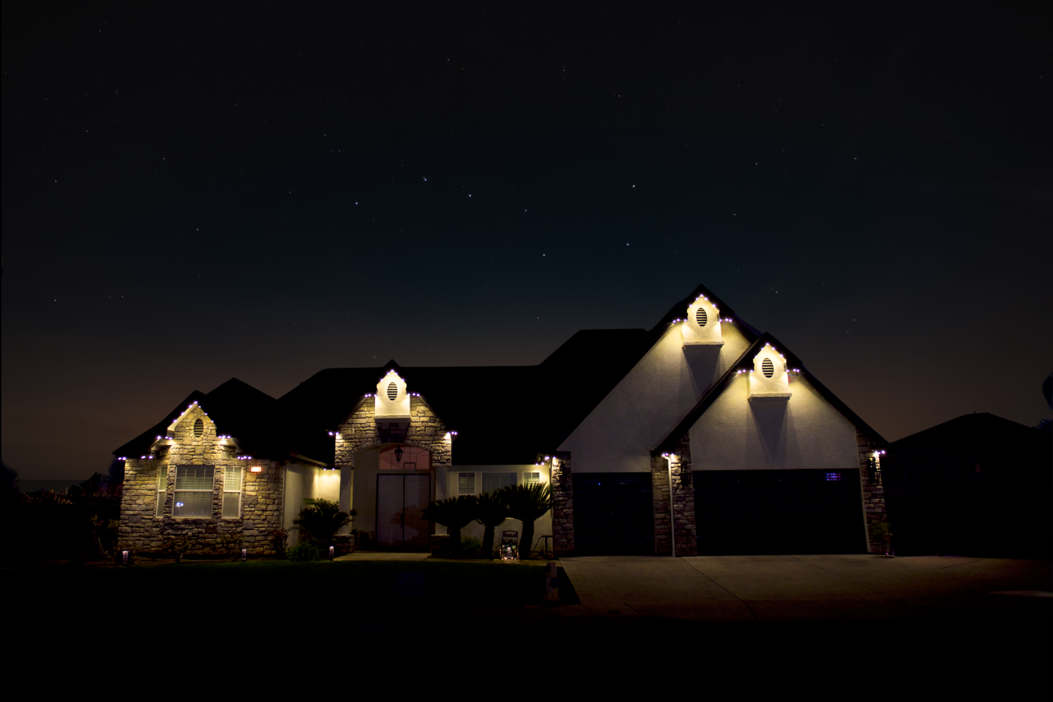 Nighttime view of a house with decorated rooflines and windows illuminated by exterior lights, under a starry sky.
