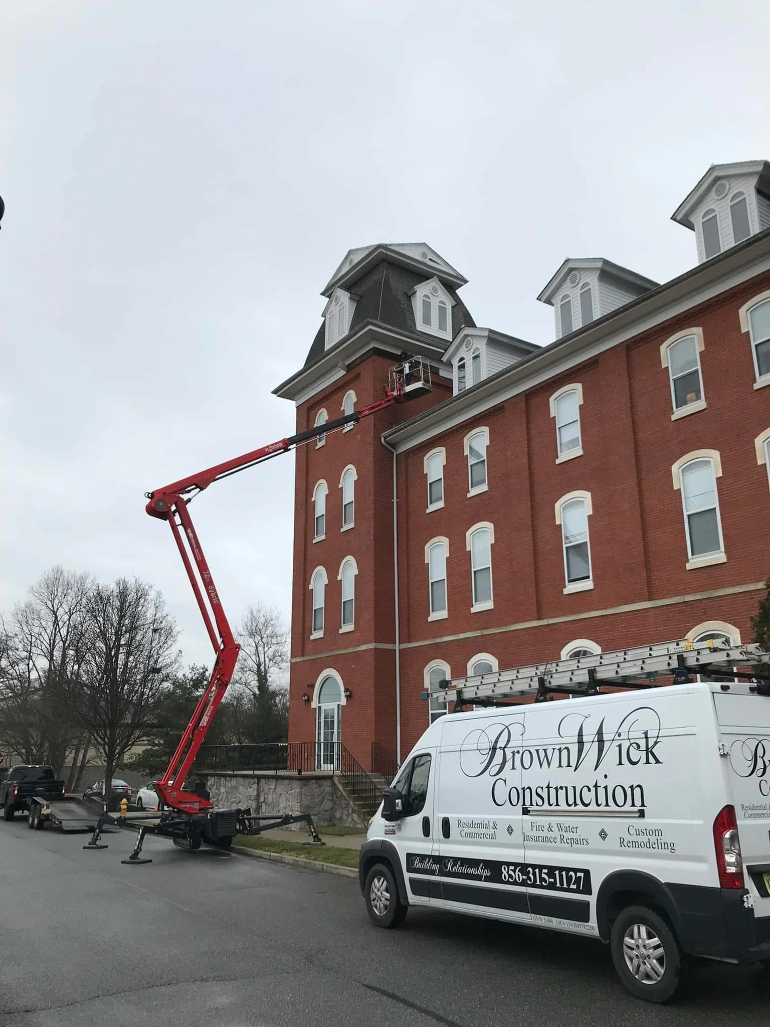A red construction lift truck is raising a worker to the top of a red brick building with white trim and multiple windows, including a tower with a dark roof. A white company van with black lettering reads 'Brown Wick Construction,' indicating the co