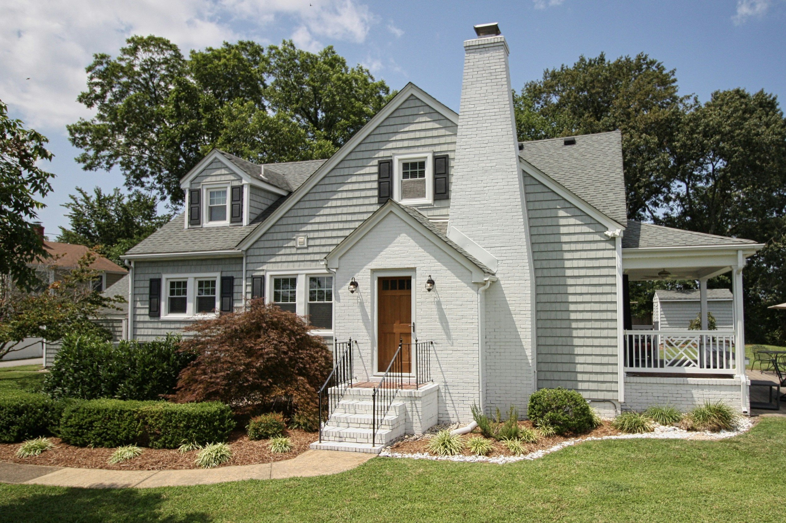 A white house with gray shingle siding and black shutters on the windows. It has a brick chimney and a front door with steps leading up to it. The house is surrounded by a well-maintained lawn, bushes, and trees under a partly cloudy sky.