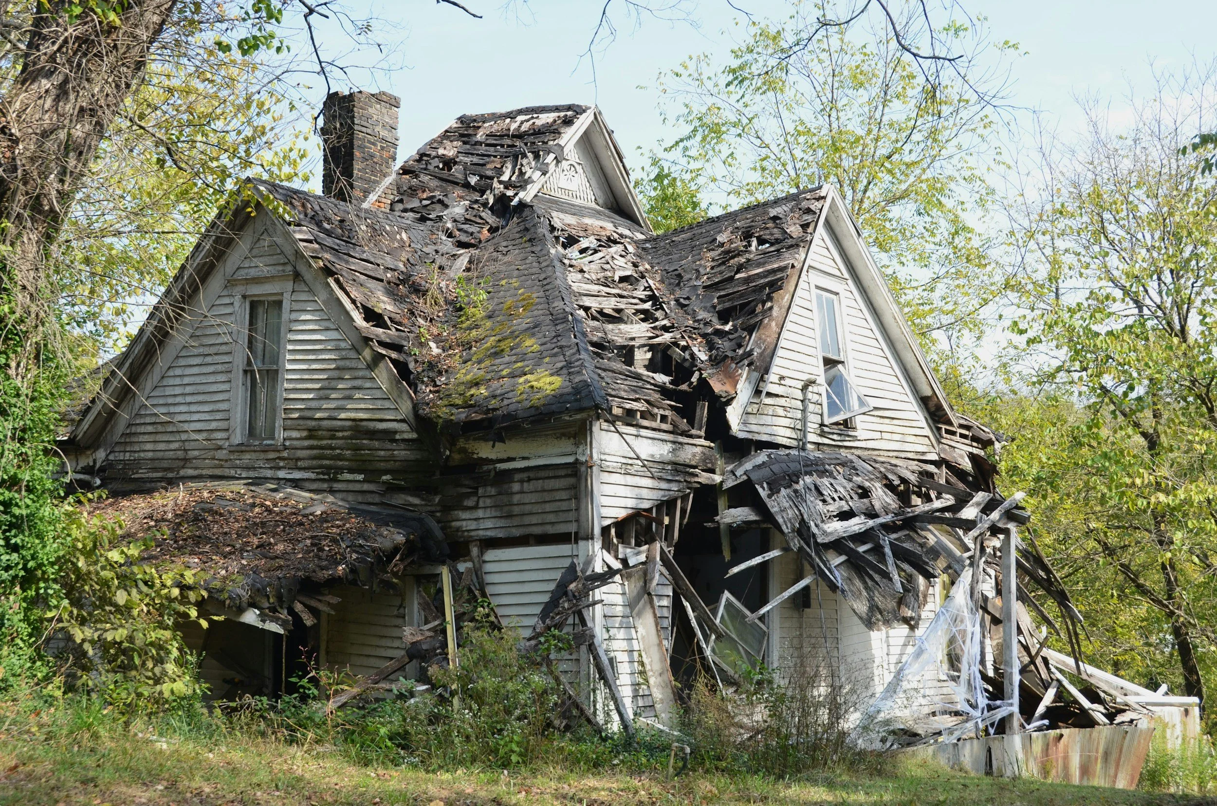 A severely damaged house with a collapsed roof and broken windows, surrounded by overgrown vegetation and trees.