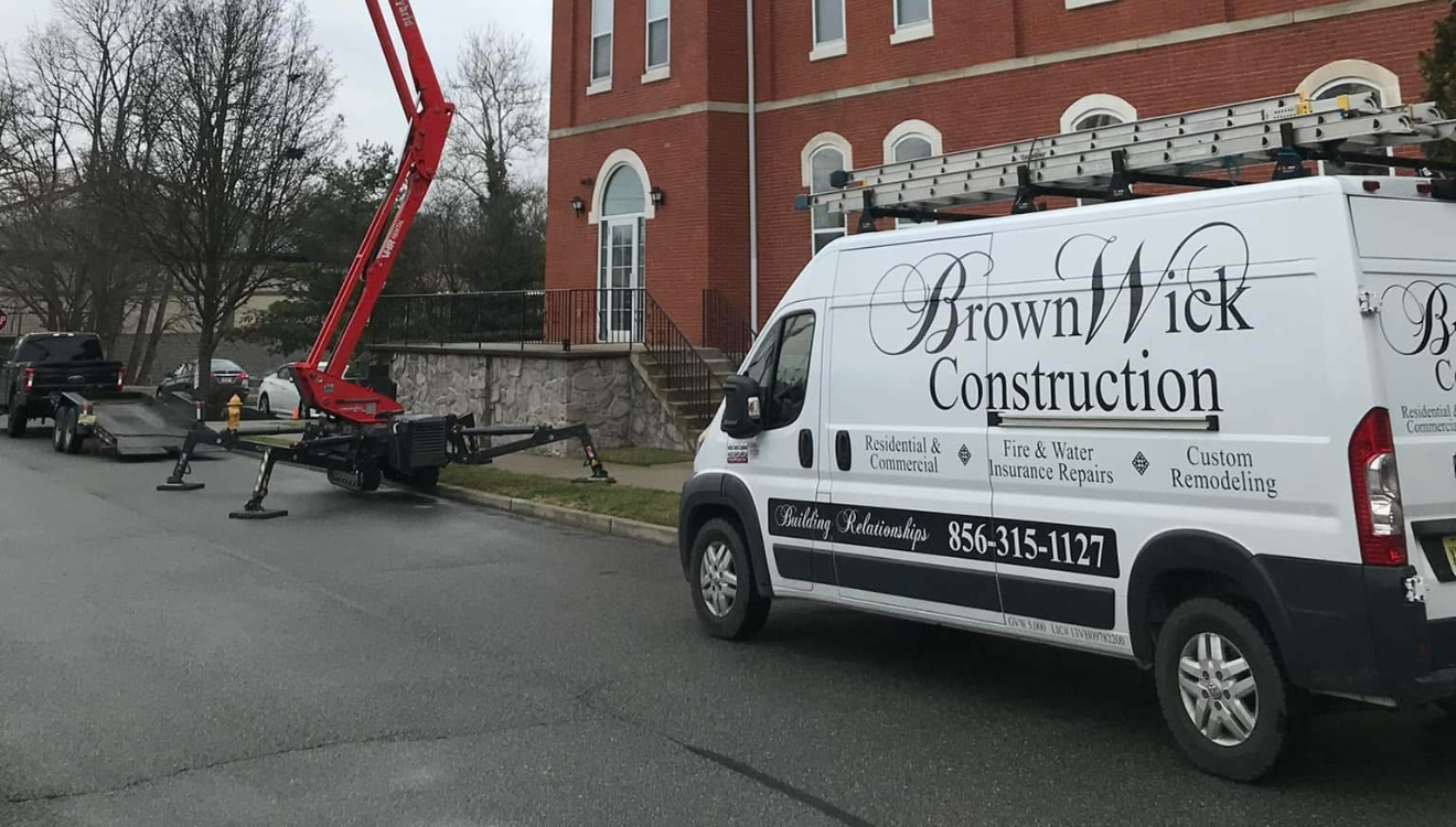 A construction vehicle with a crane arm parked on the street in front of a brick building, with a white construction van labeled "Brown Wick Construction" nearby.
