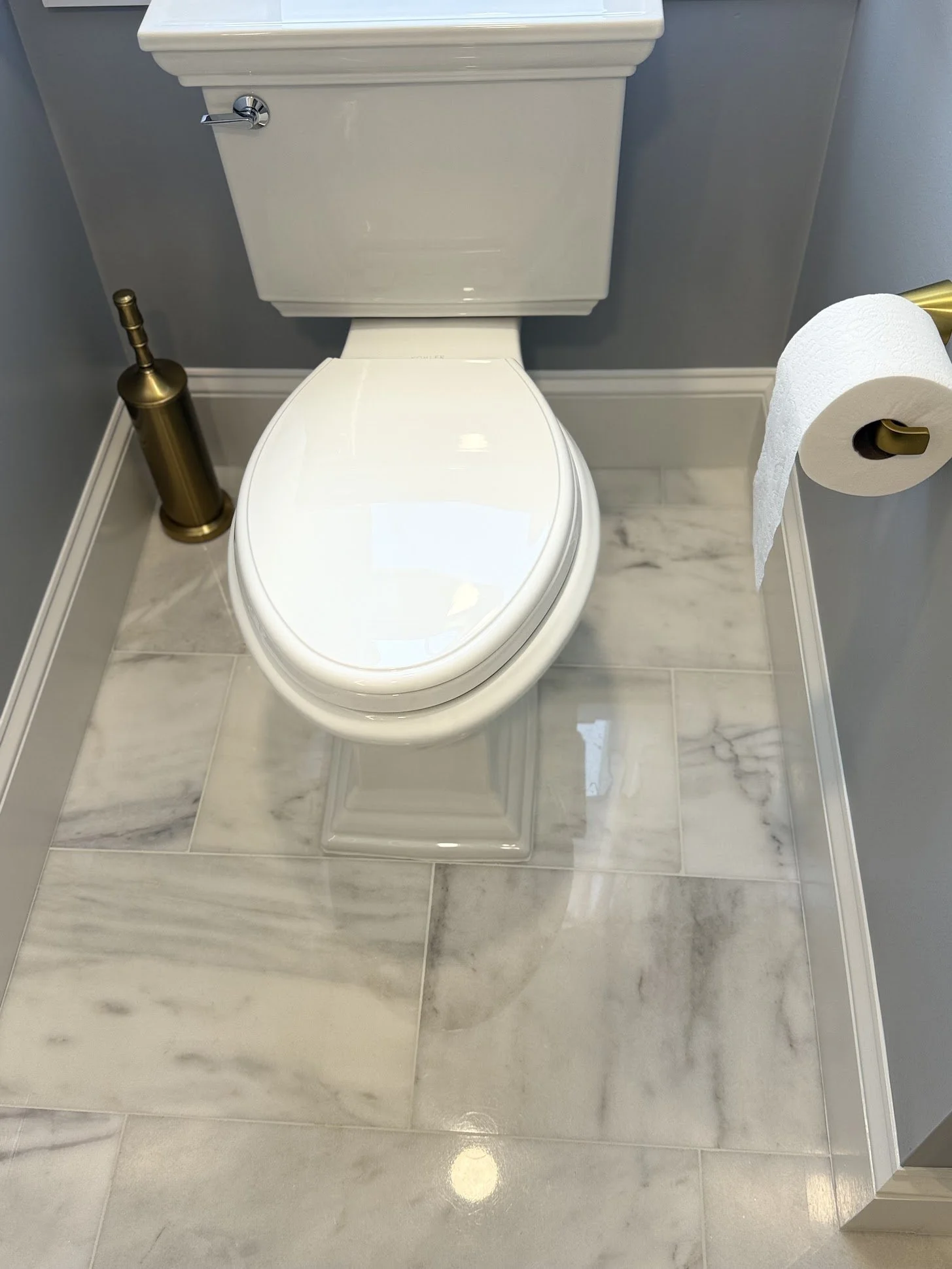 A white toilet with a closed lid, a gold toilet paper holder with a roll of toilet paper, and a gold toilet brush holder on a marble floor. The wall behind is painted light blue.