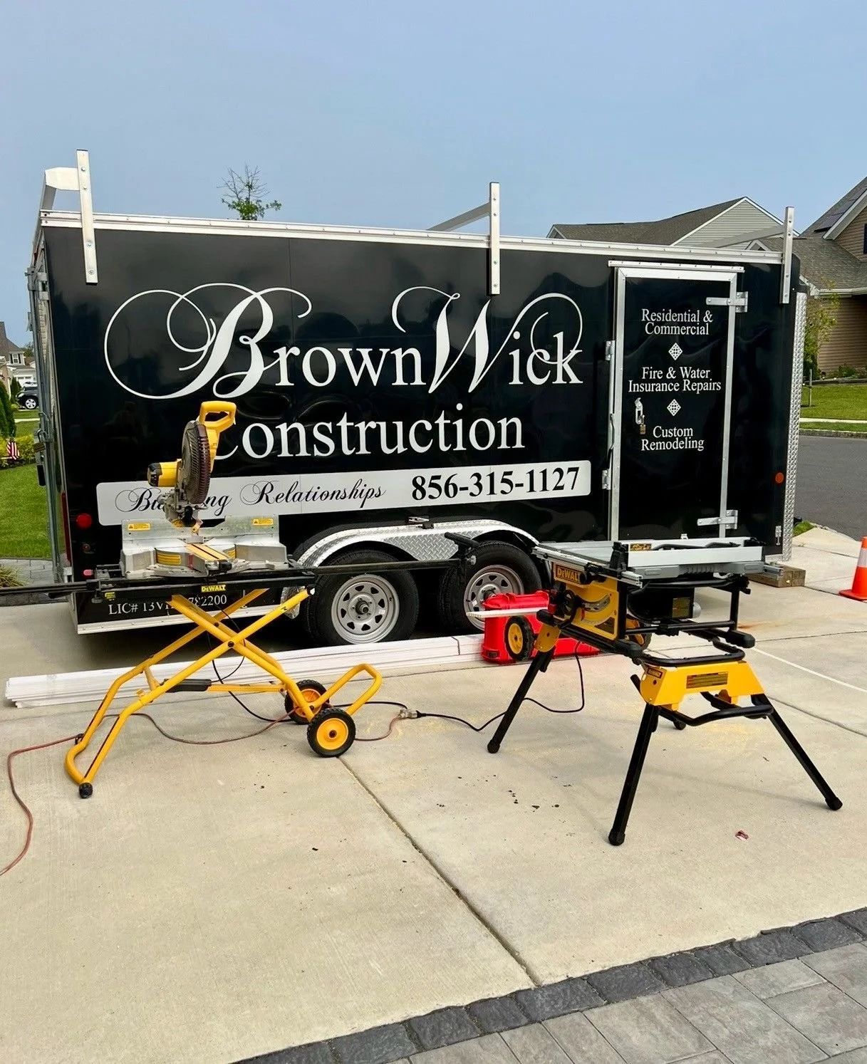 A black construction trailer with white and gold lettering advertising Brown Wick Construction, a residential and commercial construction company. The trailer is parked on a driveway with construction tools and equipment nearby, including a yellow saw, a table saw, and a cutting station. The background shows a suburban neighborhood with houses and a lawn.