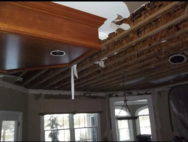 Ceiling with exposed wooden beams and incomplete drywall, with visible gaps and damage, in a room with windows.