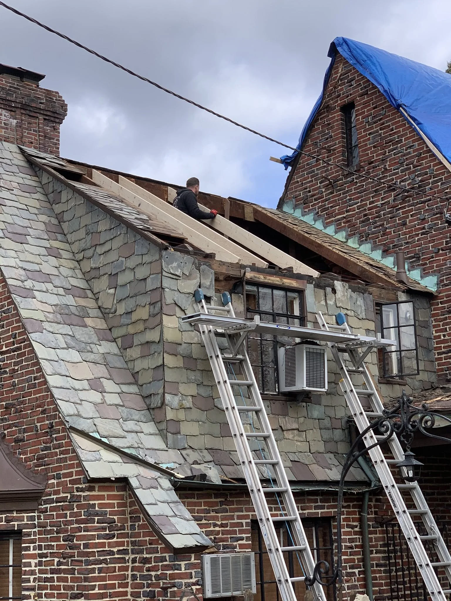A person working on the roof of a brick building, with ladders and construction materials around, and a blue tarp covering part of the roof against a cloudy sky.