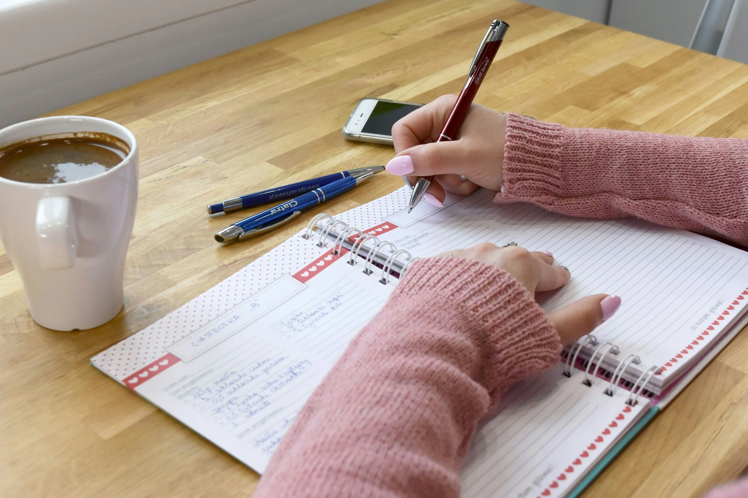 A person in a pink sweater writing in a planner with a red pen on a wooden table. There is a blue pen, a smartphone, and a white mug of coffee nearby.