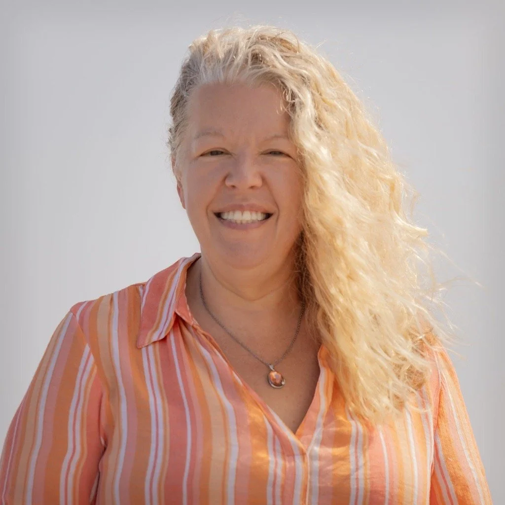 A woman with blonde, curly hair smiling outdoors in daylight, wearing a striped orange and white shirt and a necklace with a pendant.