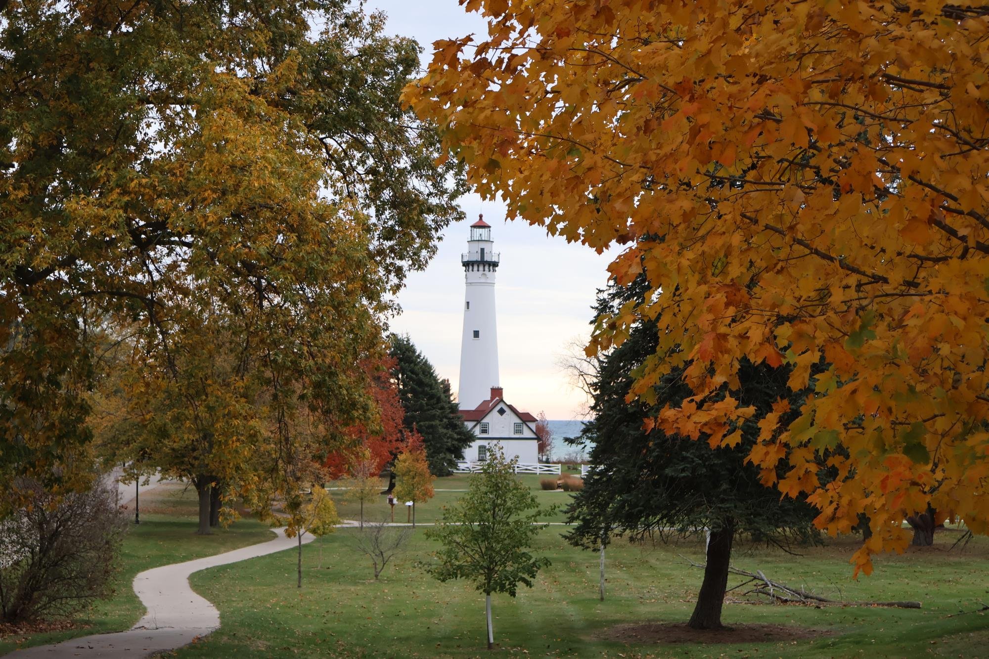 A winding pathway through a park with green grass, trees with autumn leaves, and a white lighthouse in the background.