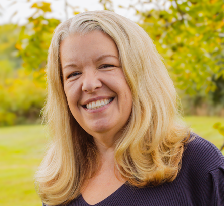 Smiling woman with blonde hair outdoors in a park with trees and grass in the background.