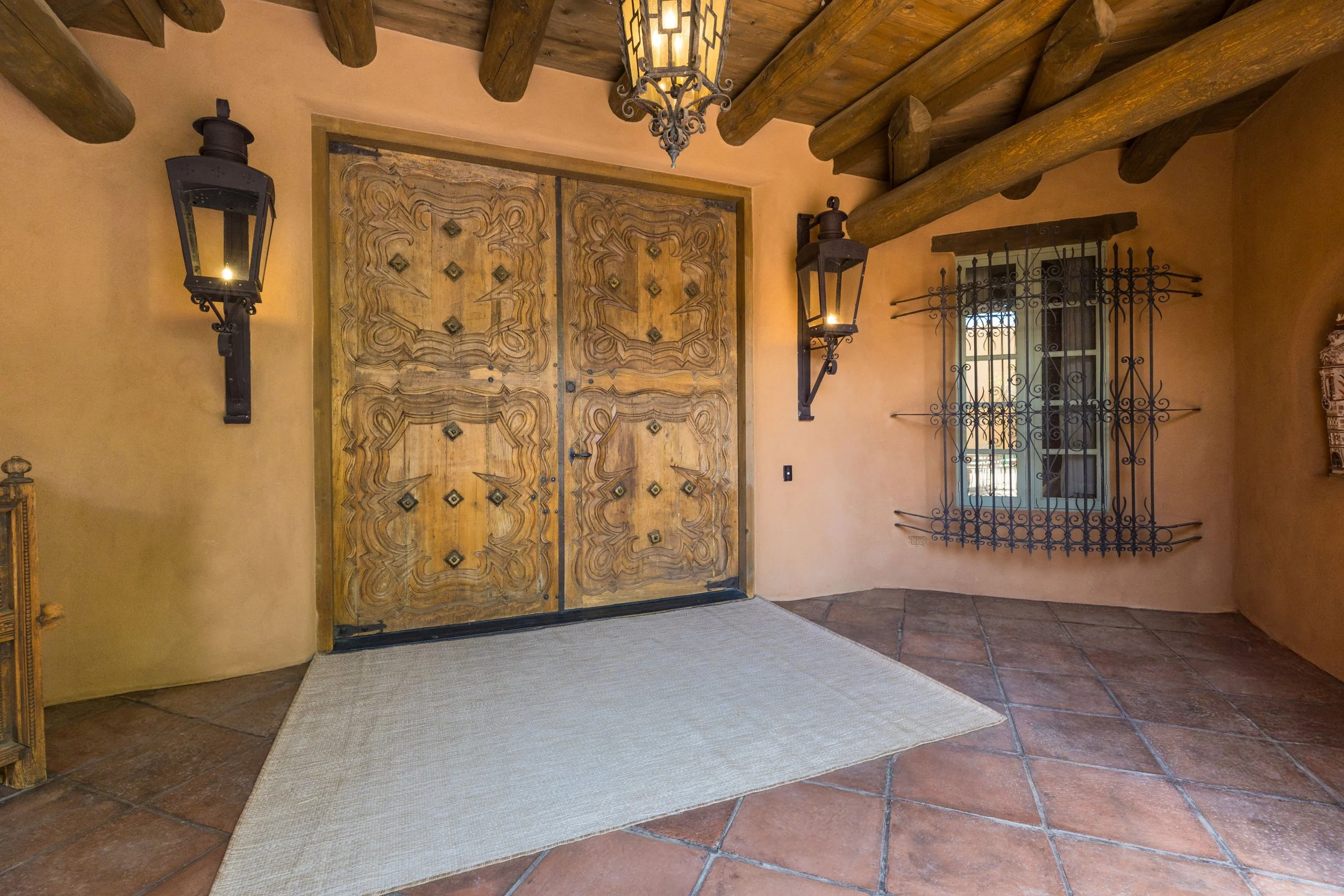 Grand zaguan entry with carved wooden double doors, lantern sconces, and Saltillo tile floors at SENDERO Taos NM — Jones West