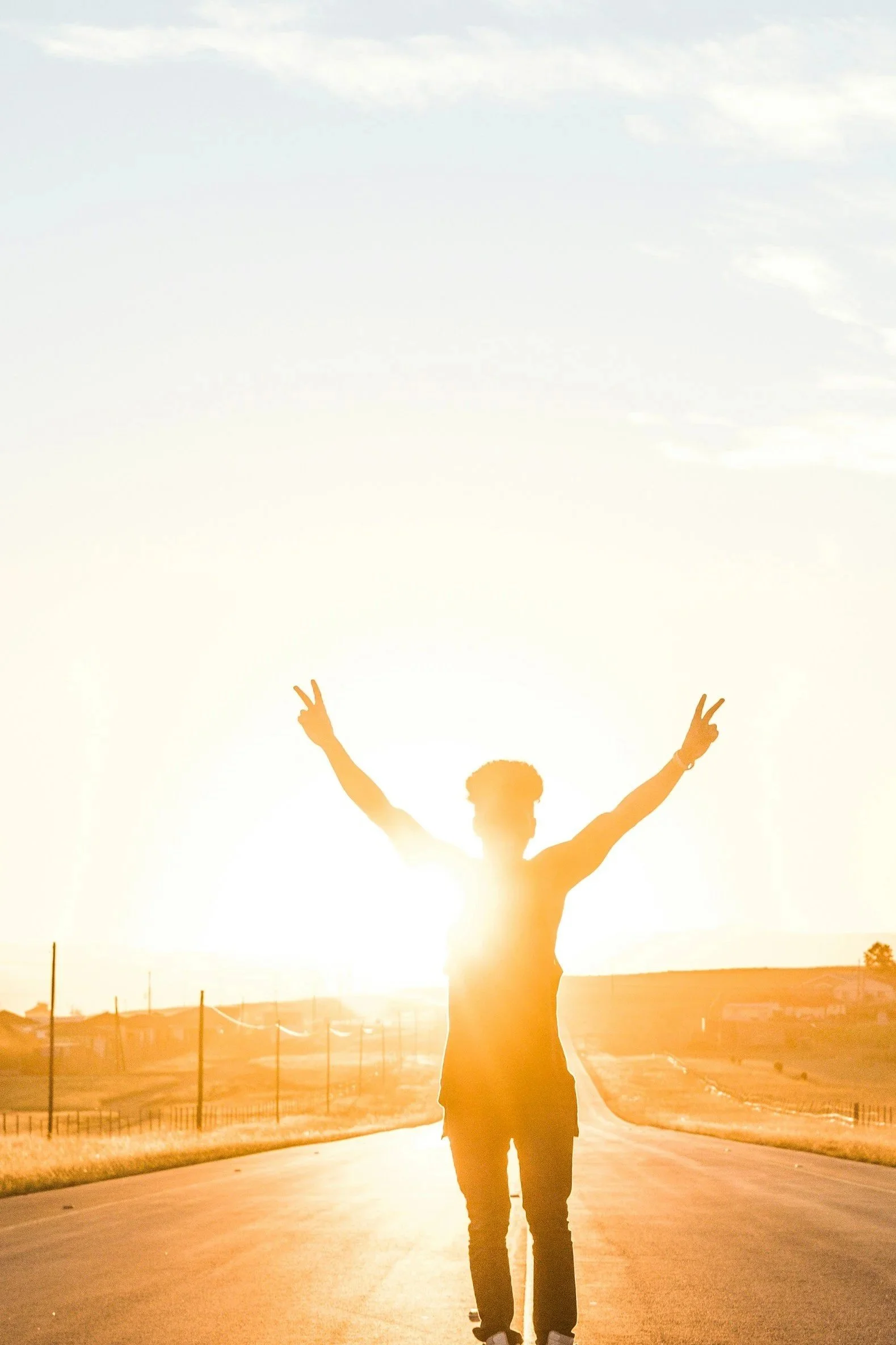 Silhouette of a person standing on a road during sunset with arms raised and making peace signs, with a clear sky and distant buildings in the background.
