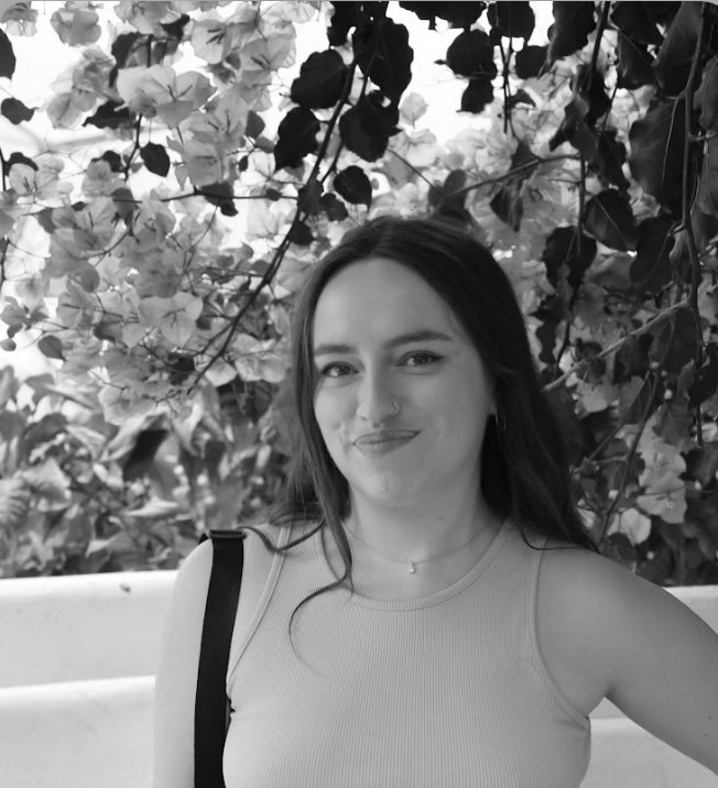 A woman with long dark hair, wearing a sleeveless top and jewelry, smiles in front of leafy foliage, in black and white.