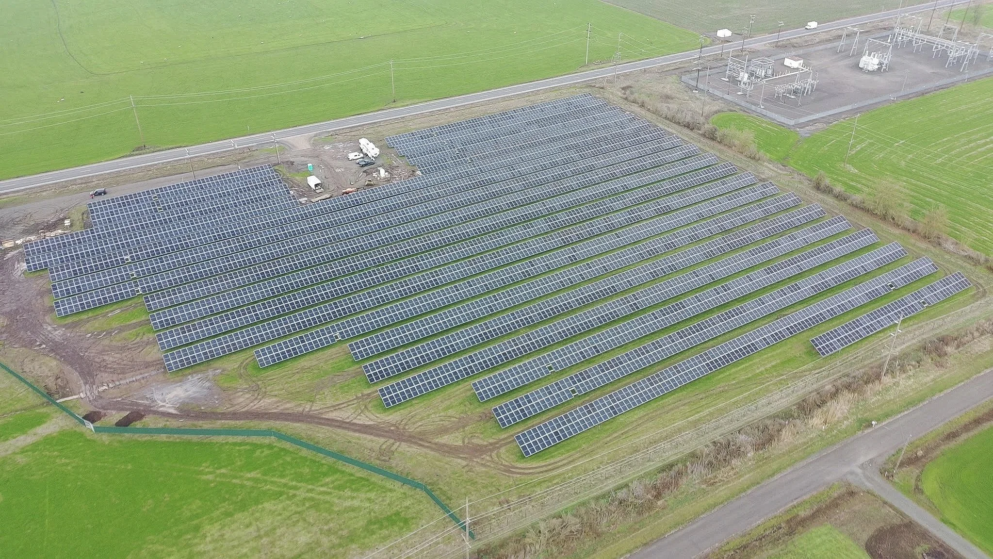 Aerial view of a large solar panel farm surrounded by green fields, with power lines and utility structures nearby.