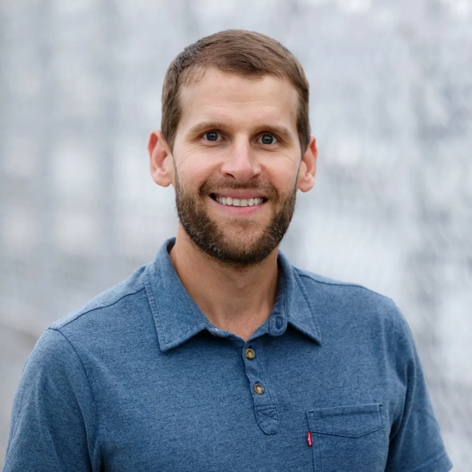 A smiling man with light skin, a beard, and blue eyes, wearing a blue button-up shirt, standing outdoors with a blurred gray background.