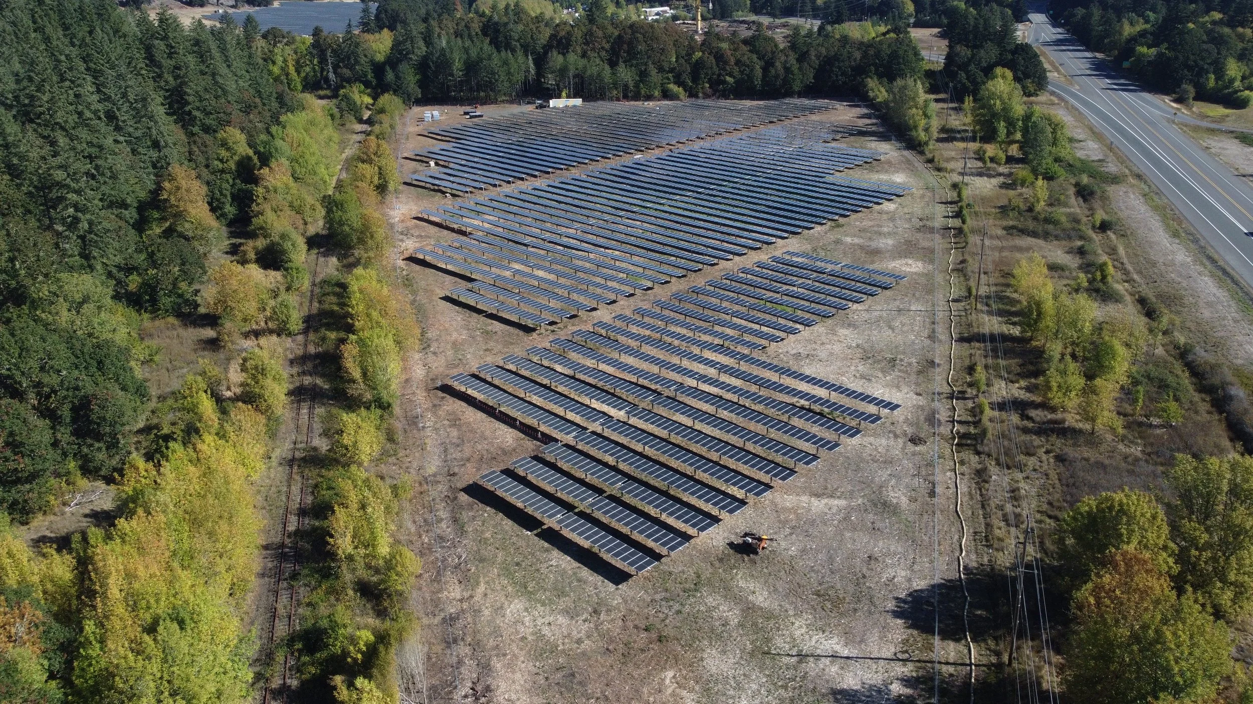 An aerial view of a large solar panel farm situated on a cleared plot of land, bordered by trees on one side and a road with power lines on the other.