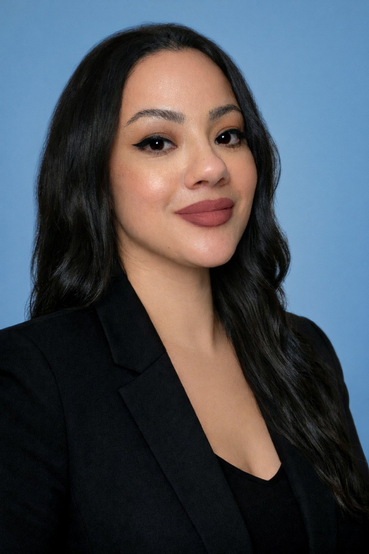 Close-up of a woman with long black hair, wearing a black blazer and lipstick, standing against a blue background.