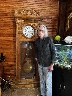 Man with glasses standing beside a tall wooden grandfather clock in a room with wooden walls and a fish tank.