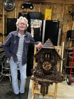 An older man with gray hair and glasses standing in a garage, leaning on a large decorative cuckoo clock. The garage has wooden walls, a refrigerator covered with magnets, and various tools and items stored around.