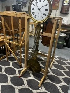 Floor clock with a large round face and Roman numerals, standing on a patterned rug in a store with wooden furniture and other clocks in the background.