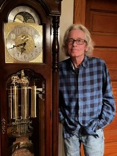 Older man with gray hair and glasses standing next to a tall wooden grandfather clock.